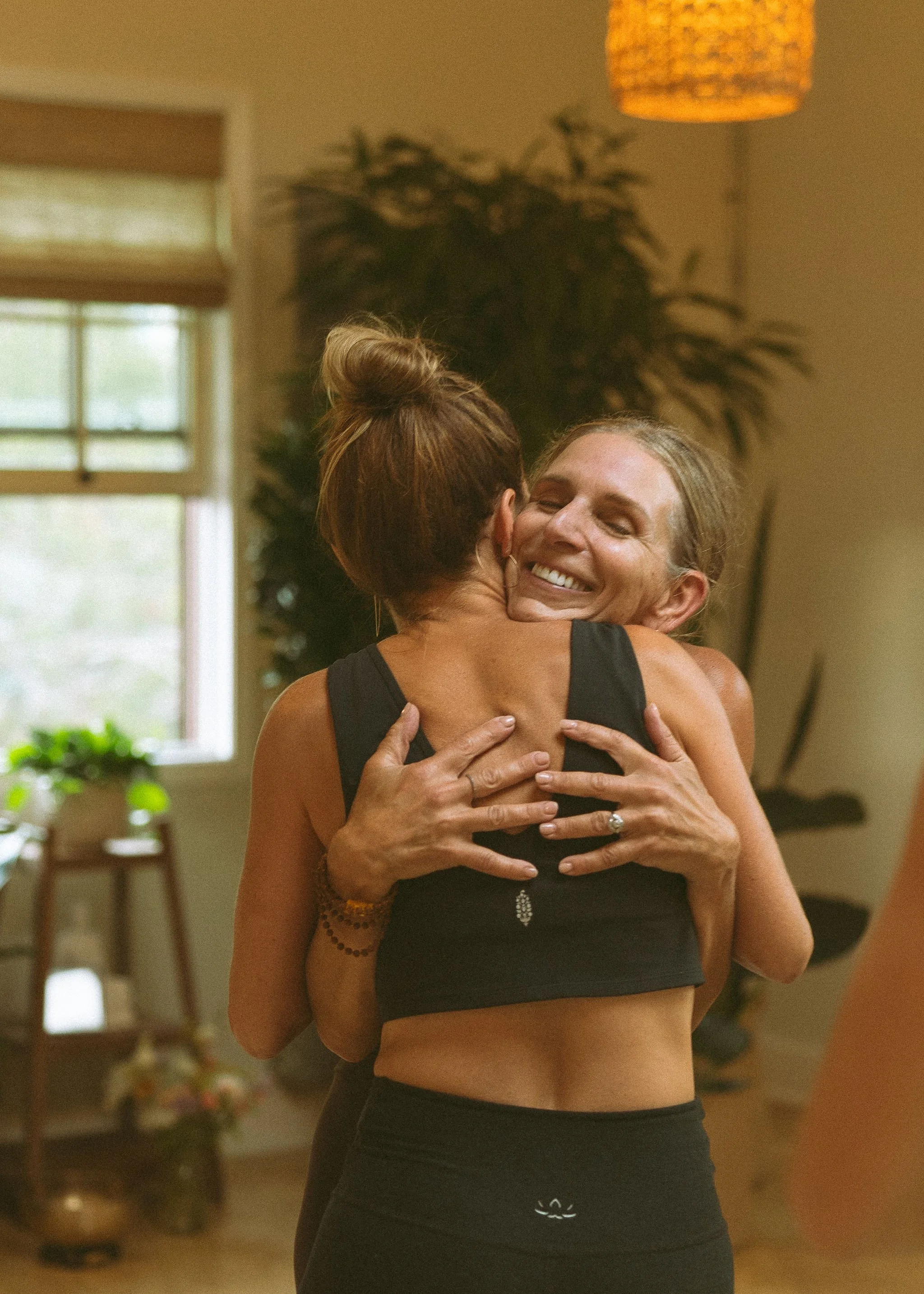 Two women hugging in a room with a window and warm lighting, one with gray hair and the other with brown hair in a bun.