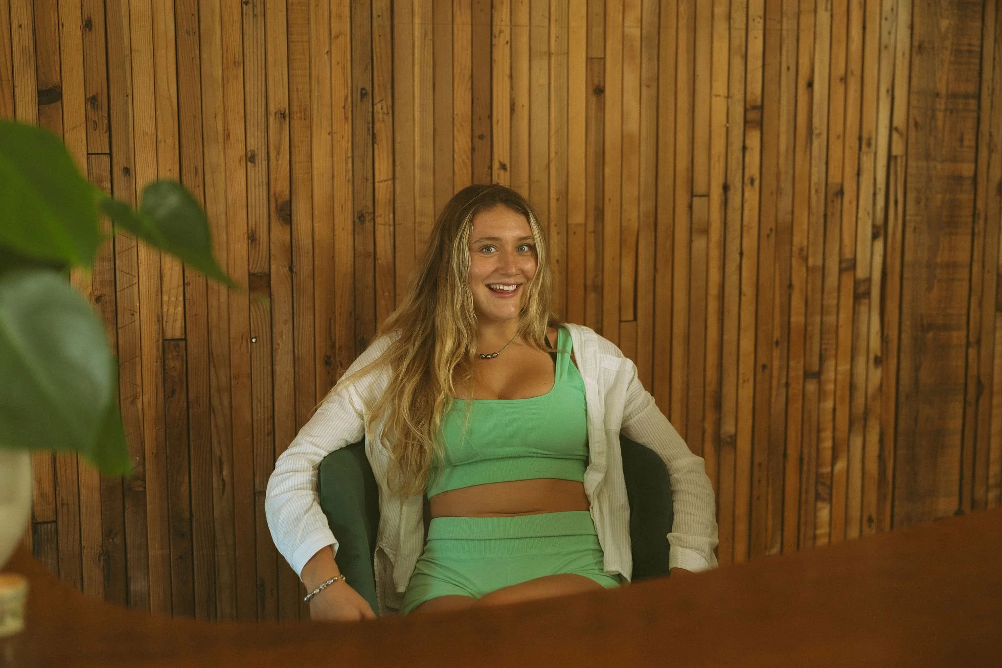 A young woman with long blonde hair, wearing a green sleeveless top and a white jacket, sitting on a black chair in front of a wooden wall. She is smiling and looking at the camera.