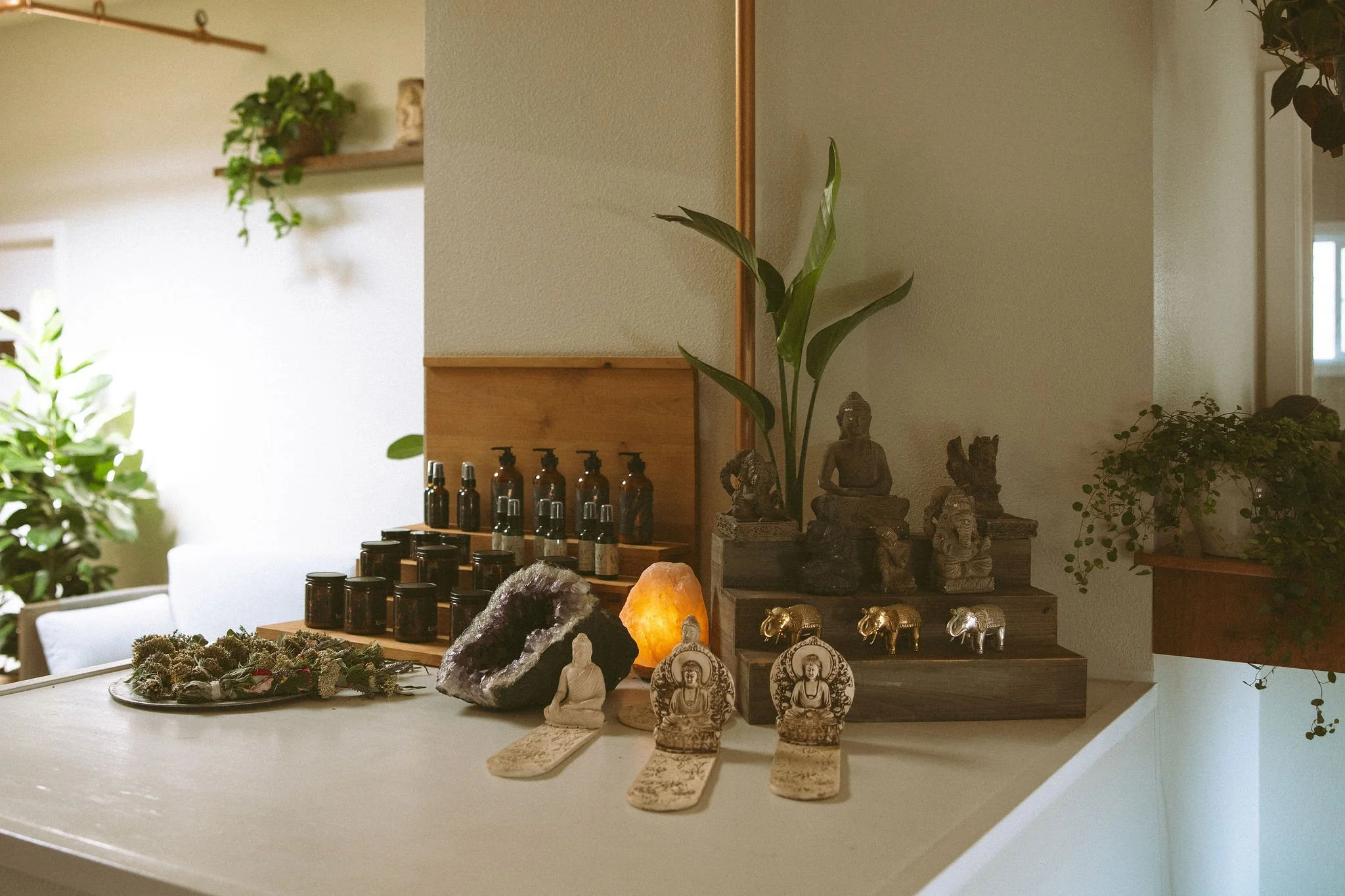 A display of various spiritual and decorative objects on a table and shelves, including small statues of Buddha, animal figurines, Himalayan salt lamp, crystals, and essential oil bottles, with green plants and wooden accents in the background.