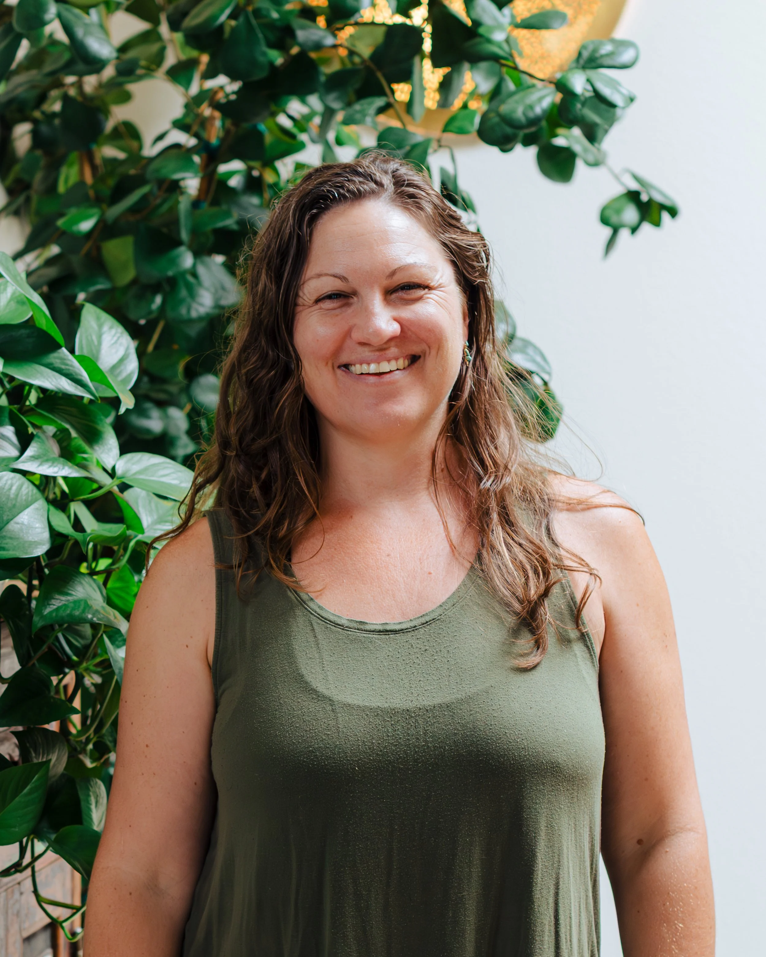 A woman smiling in front of lush green plants, wearing a green sleeveless top.