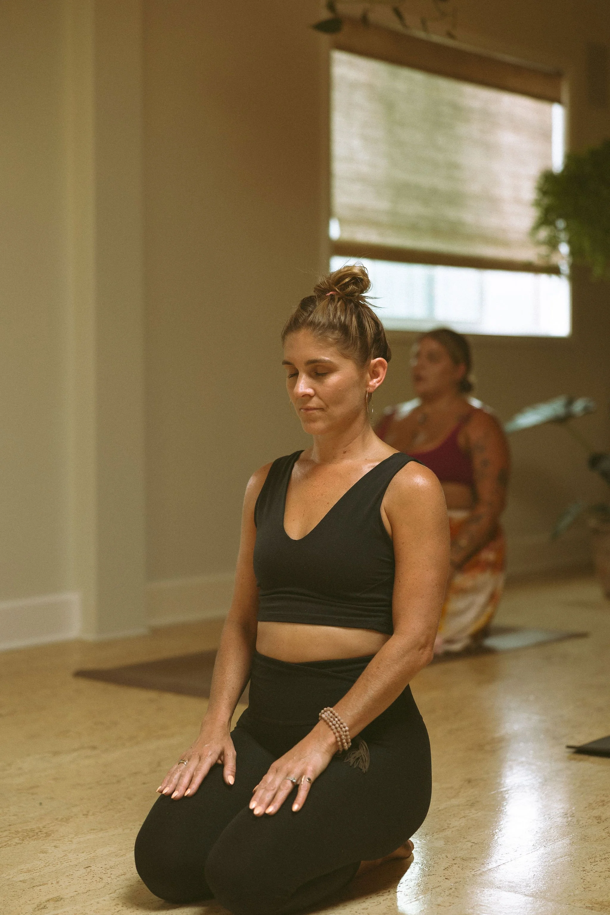 A woman in a black sports bra and leggings is kneeling with her hands resting on her thighs, eyes closed, in a yoga or meditation pose in a studio with a woman in the background.