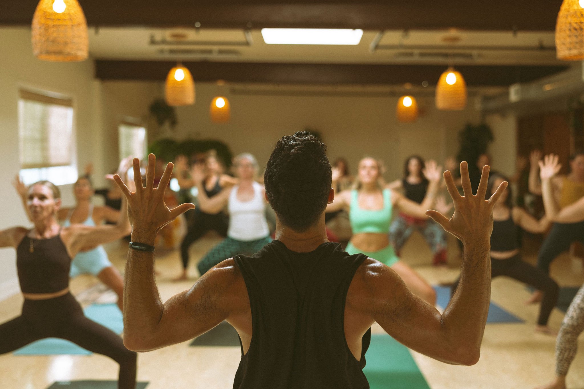 A fitness instructor leading a yoga or exercise class in a studio, with participants doing stretches or yoga poses, seen from behind.