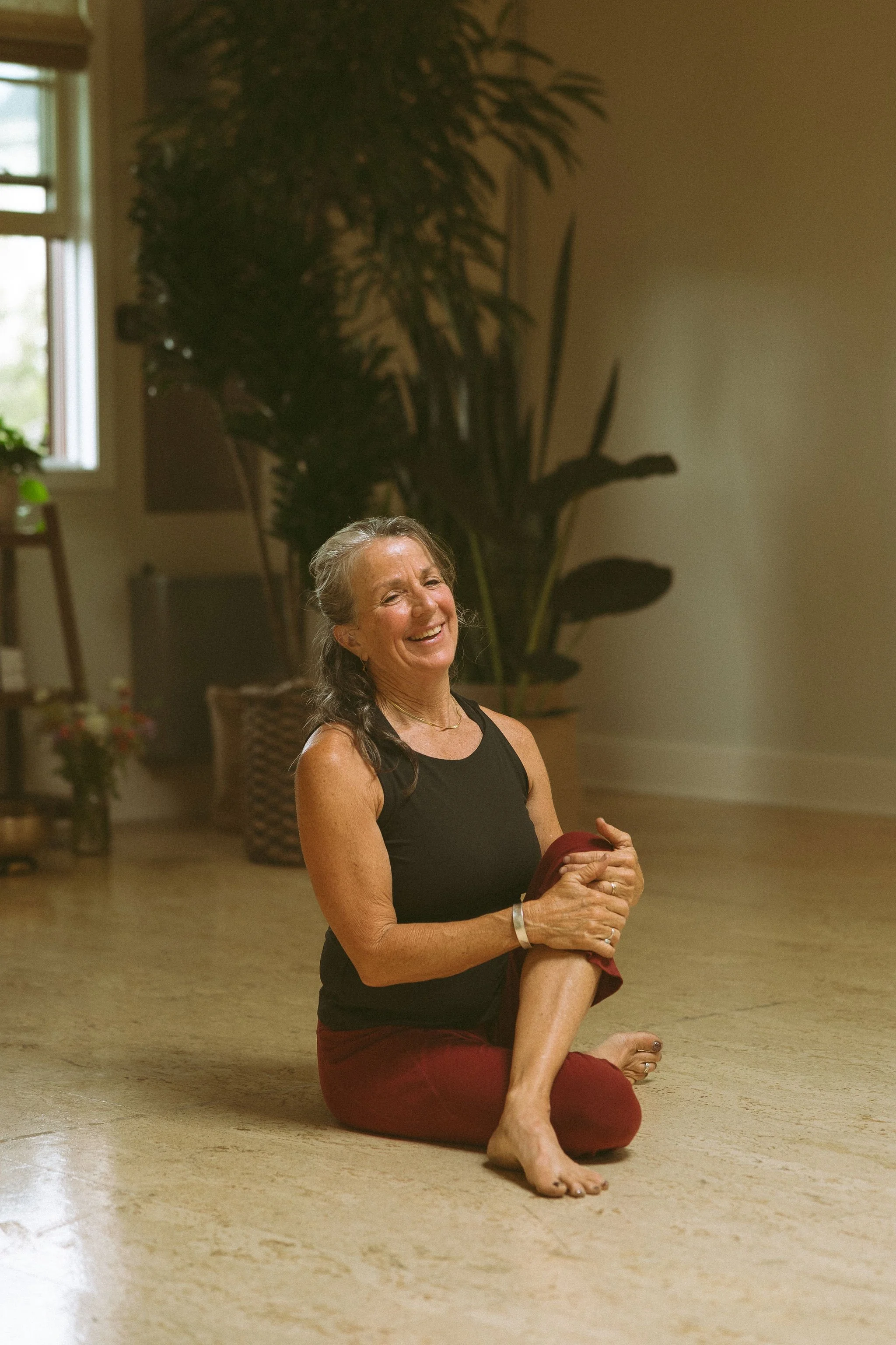A woman practicing yoga indoors, sitting on the floor with one leg bent and the other leg extended, holding her foot while smiling. She is wearing a black tank top and red pants, with a large plant and window in the background.