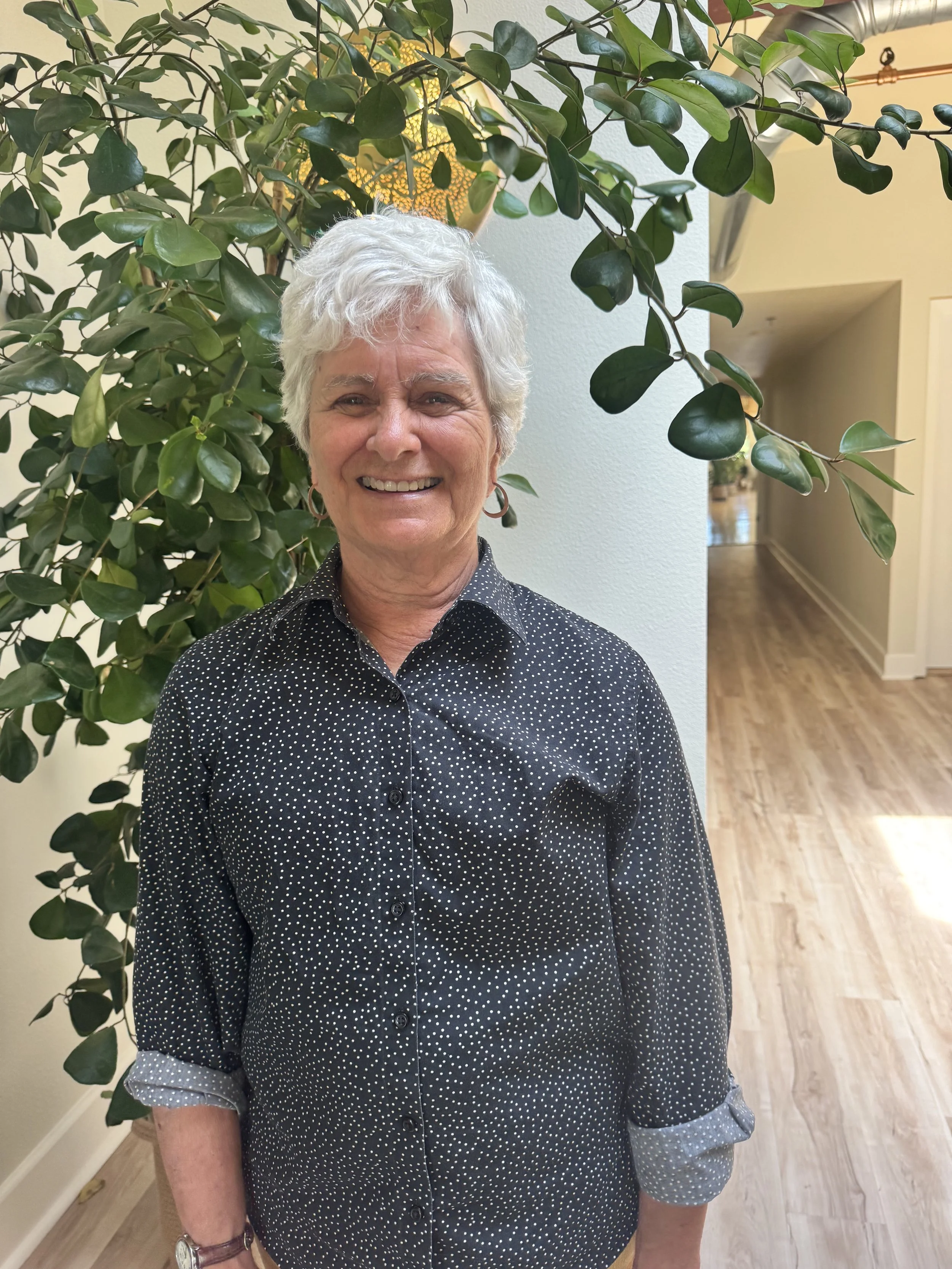 A woman with short gray hair, wearing a black shirt with white polka dots, standing inside near a large green leafy plant and smiling.