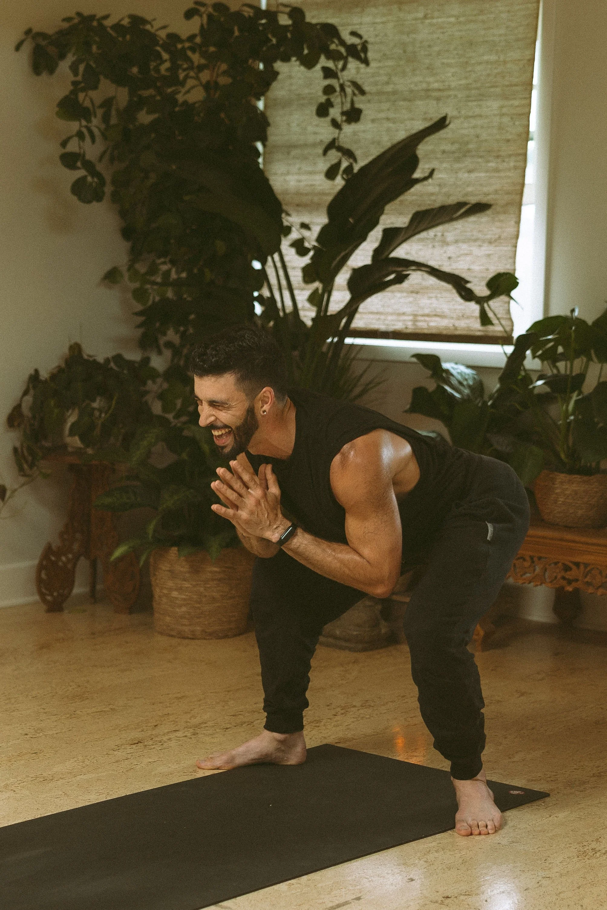A man practicing yoga indoors on a black mat. He is smiling, wearing a black sleeveless shirt and black pants, and is holding a pose with hands clasped together in front of his chest. The room has wooden floors, large green plants, a wicker basket, a