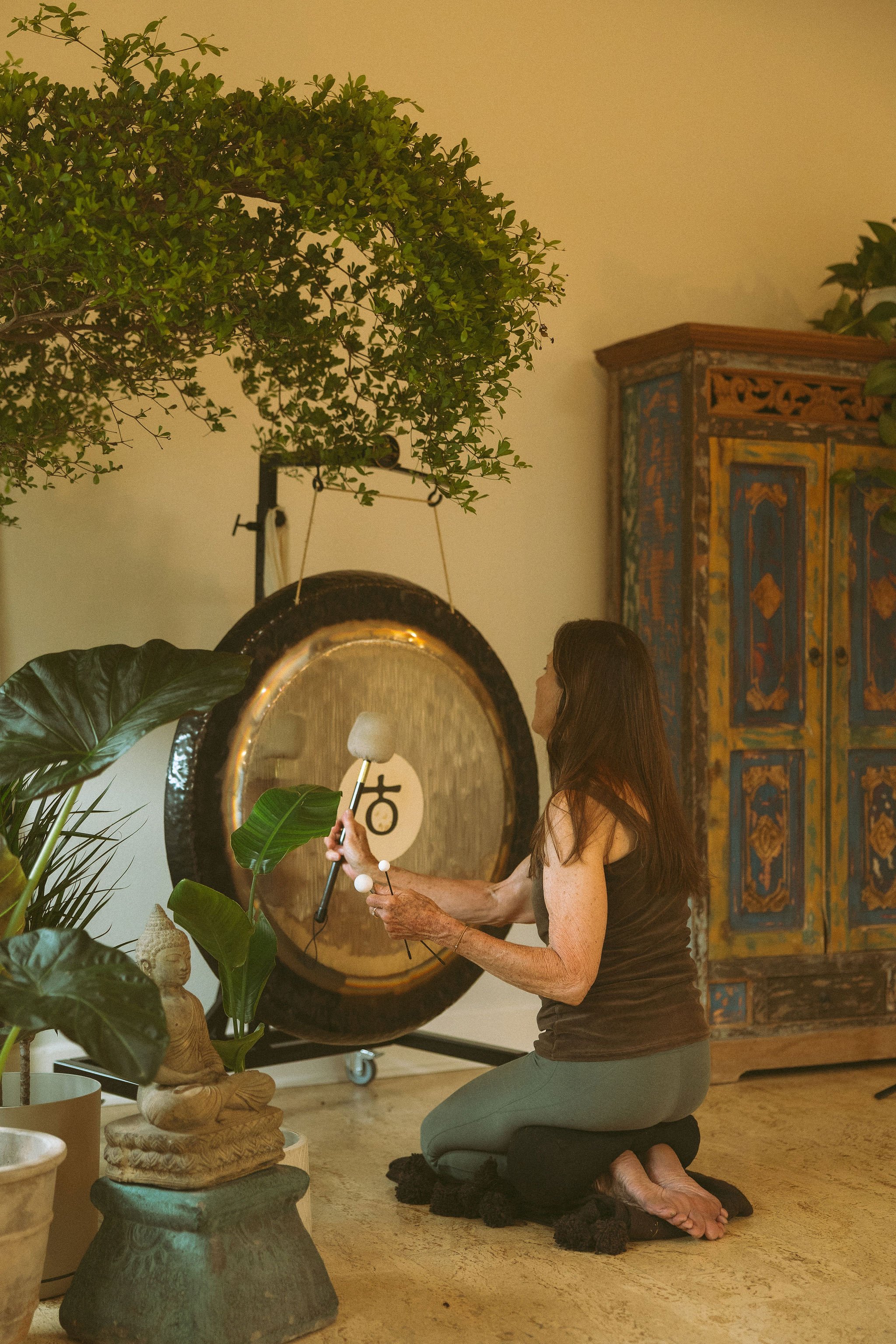An elderly woman kneeling on a black cushion, practicing meditation or mindfulness, holding a singing bowl and a stick, surrounded by plants and statues, with a large antique colorful cabinet in the background.