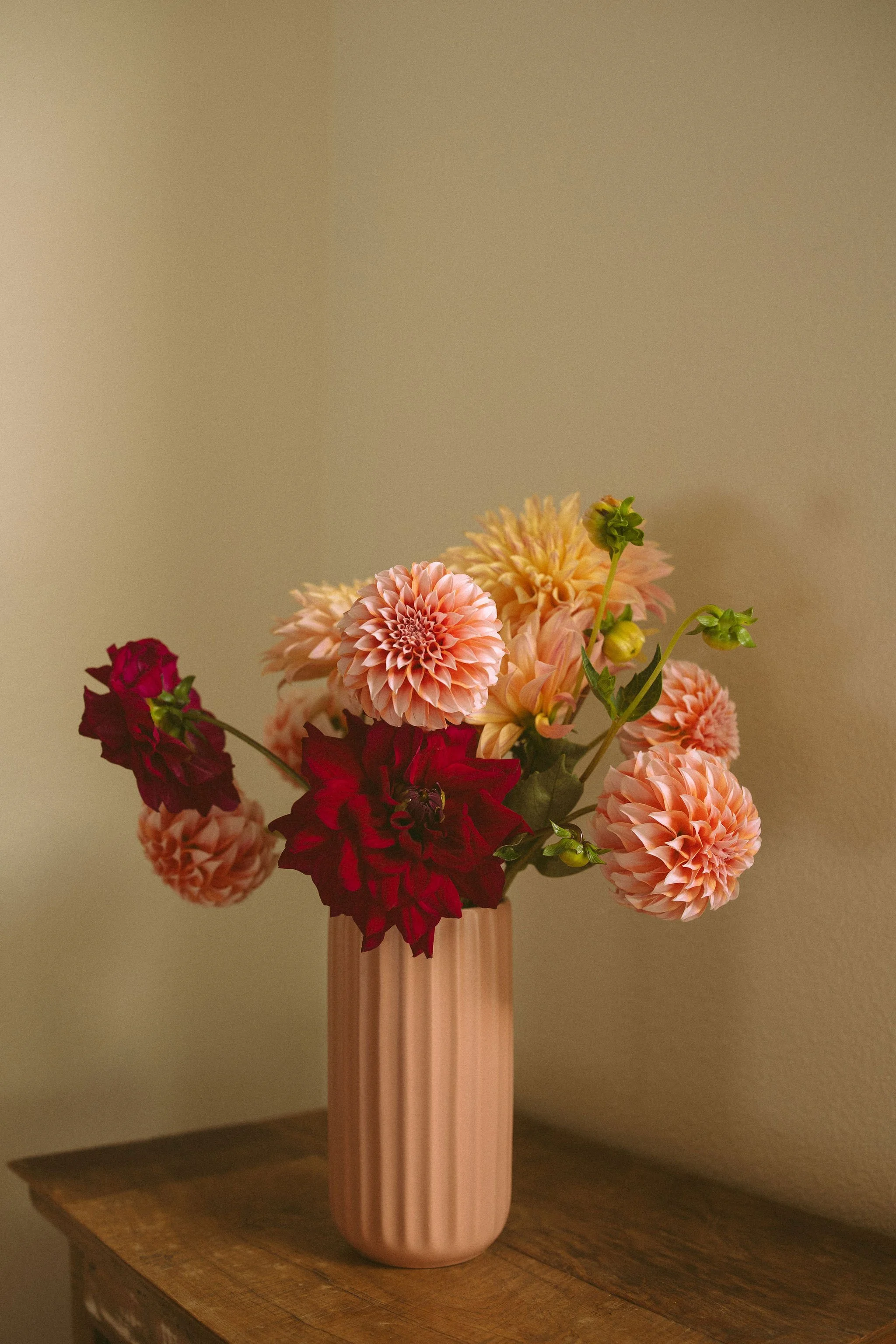 A pink vase with a bouquet of pink, red, peach, and green flowers, placed on a wooden table against a beige wall.