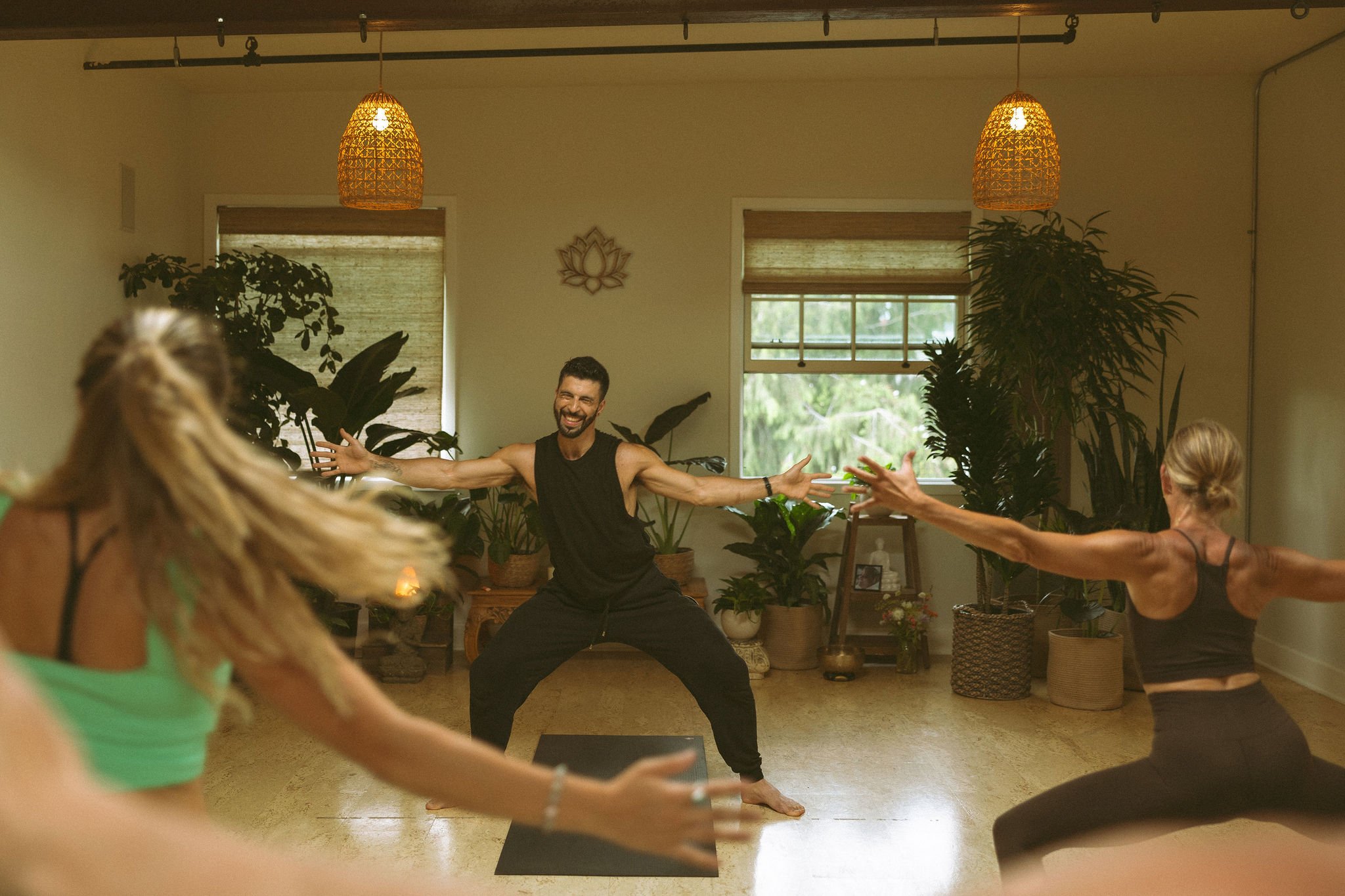 A group of people participating in a yoga or dance class indoors. A man with a beard in a black sleeveless shirt is in the center, smiling with arms outstretched. Others are partially visible, reaching towards the man. The room is decorated with plan