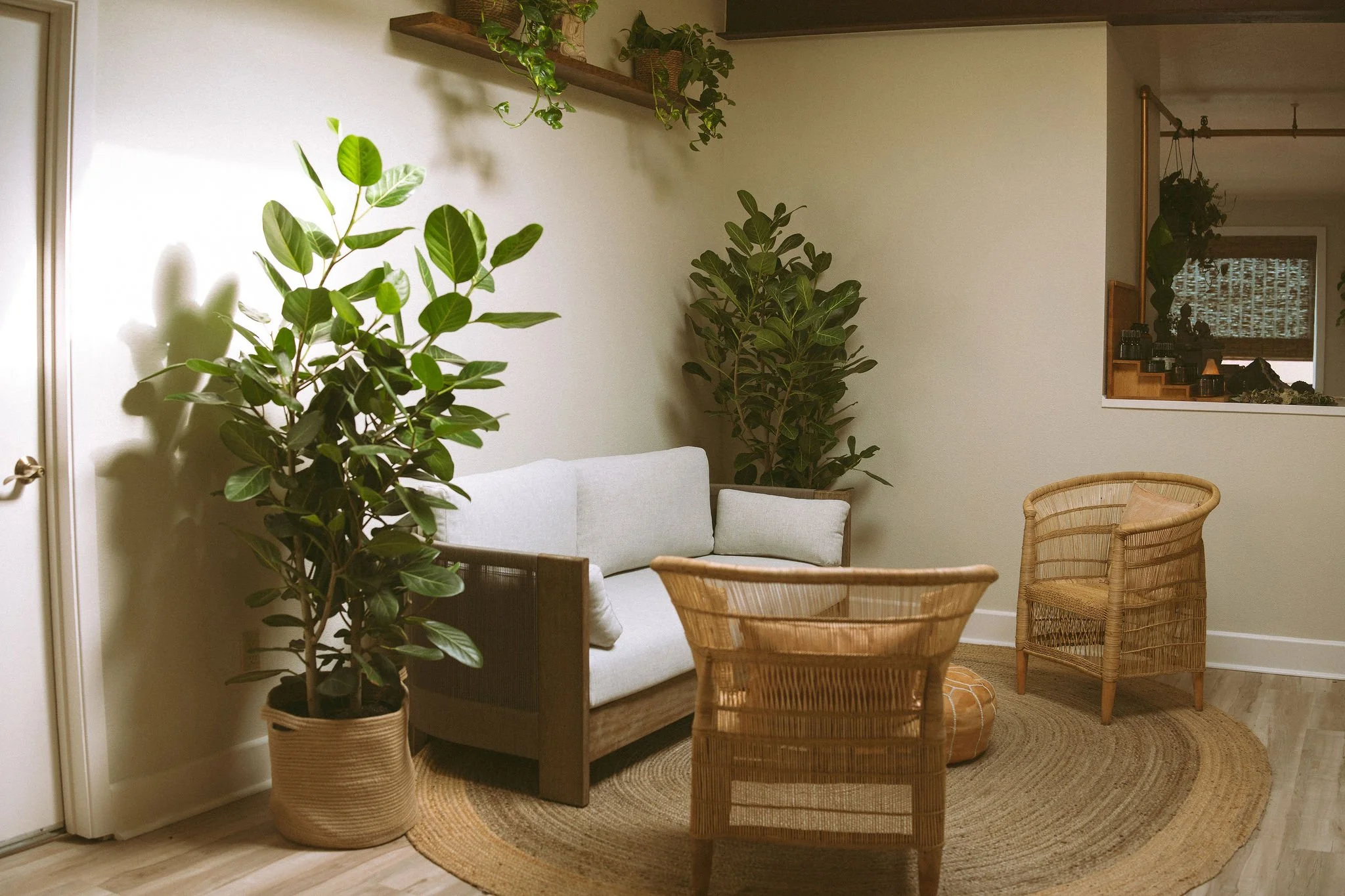 A cozy living room corner with a white sofa, wicker chairs, potted plants, a round woven rug, and a wooden shelf with more plants. There are warm tones and natural decor elements.