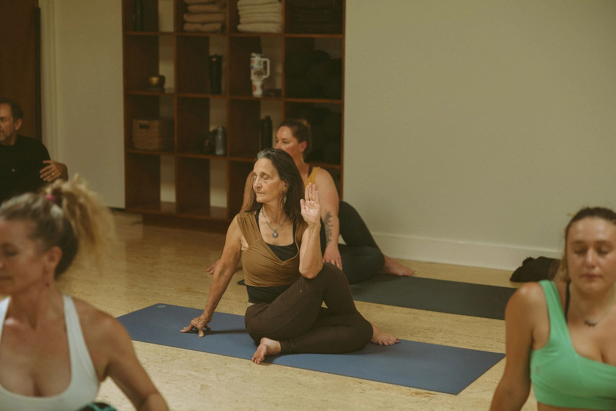 Women practicing yoga in a group class, with one woman in a kneeling pose with her hand raised and eyes closed, in a room with wooden flooring, shelves with folded towels, and a beige wall.