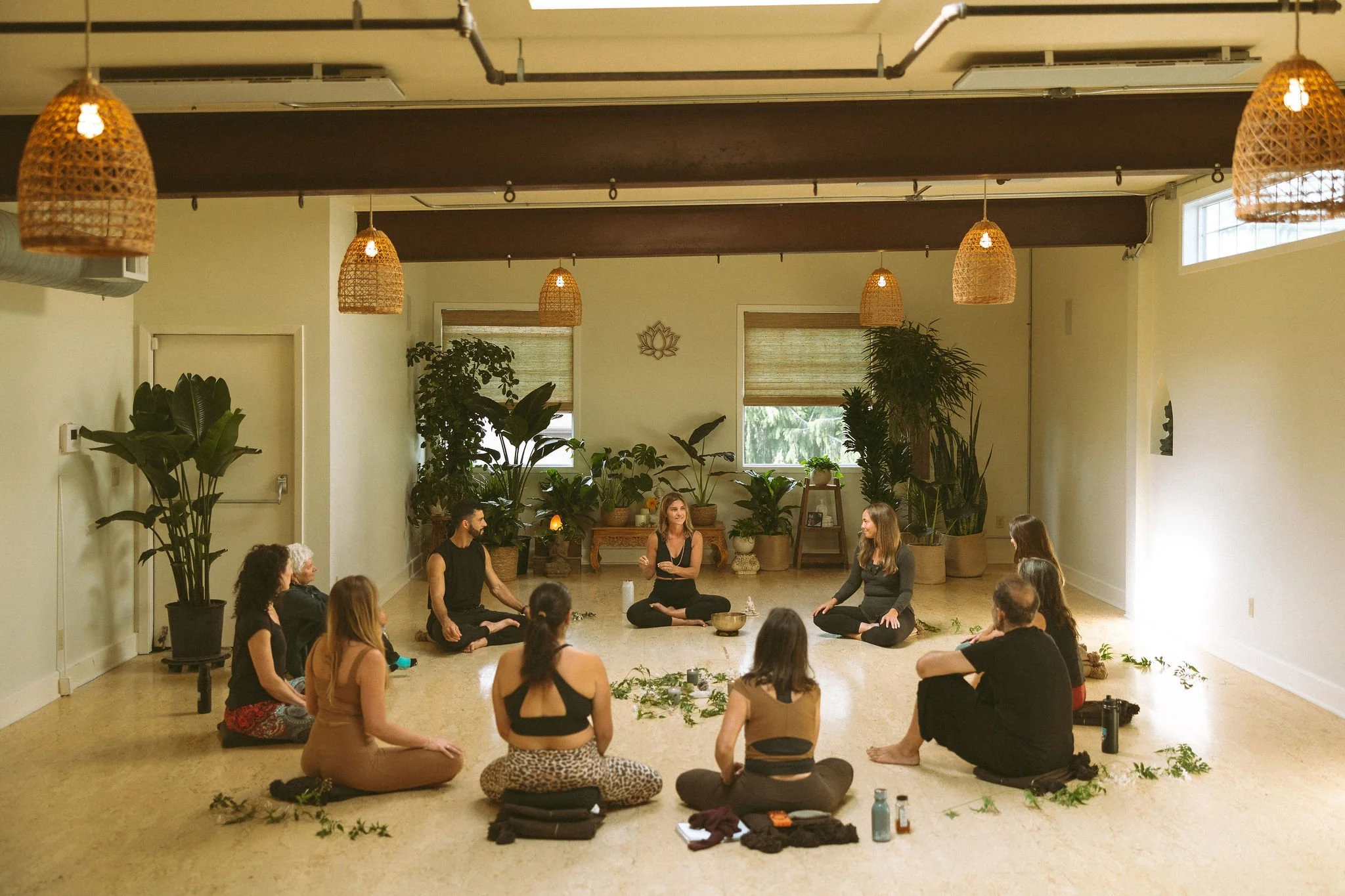 A group of people participating in a meditation or yoga session in a spacious, well-lit room with indoor plants and hanging wicker light fixtures, sitting in a circle on the floor.