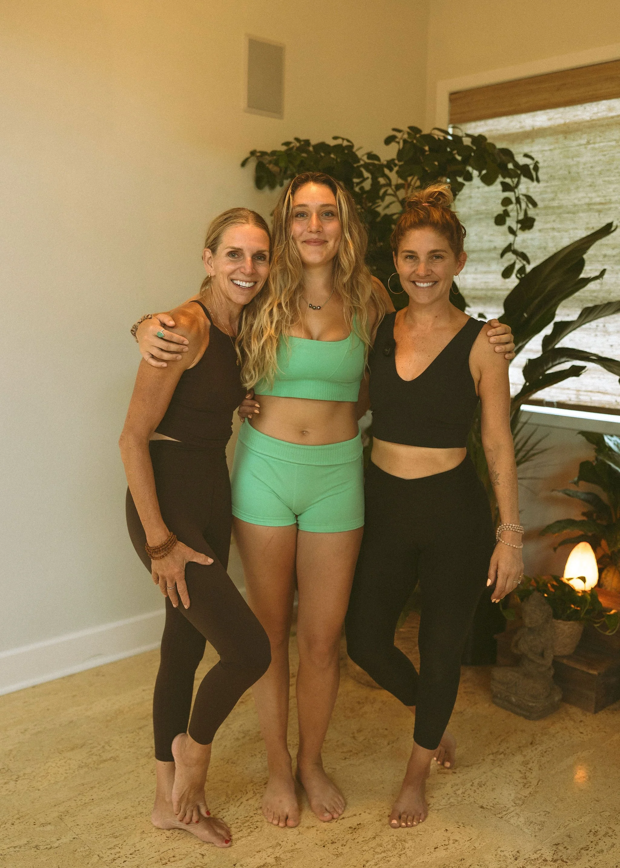 Three women standing together indoors, smiling, with one in a green workout outfit and the other two in black athletic wear, surrounded by plants and warm lighting.