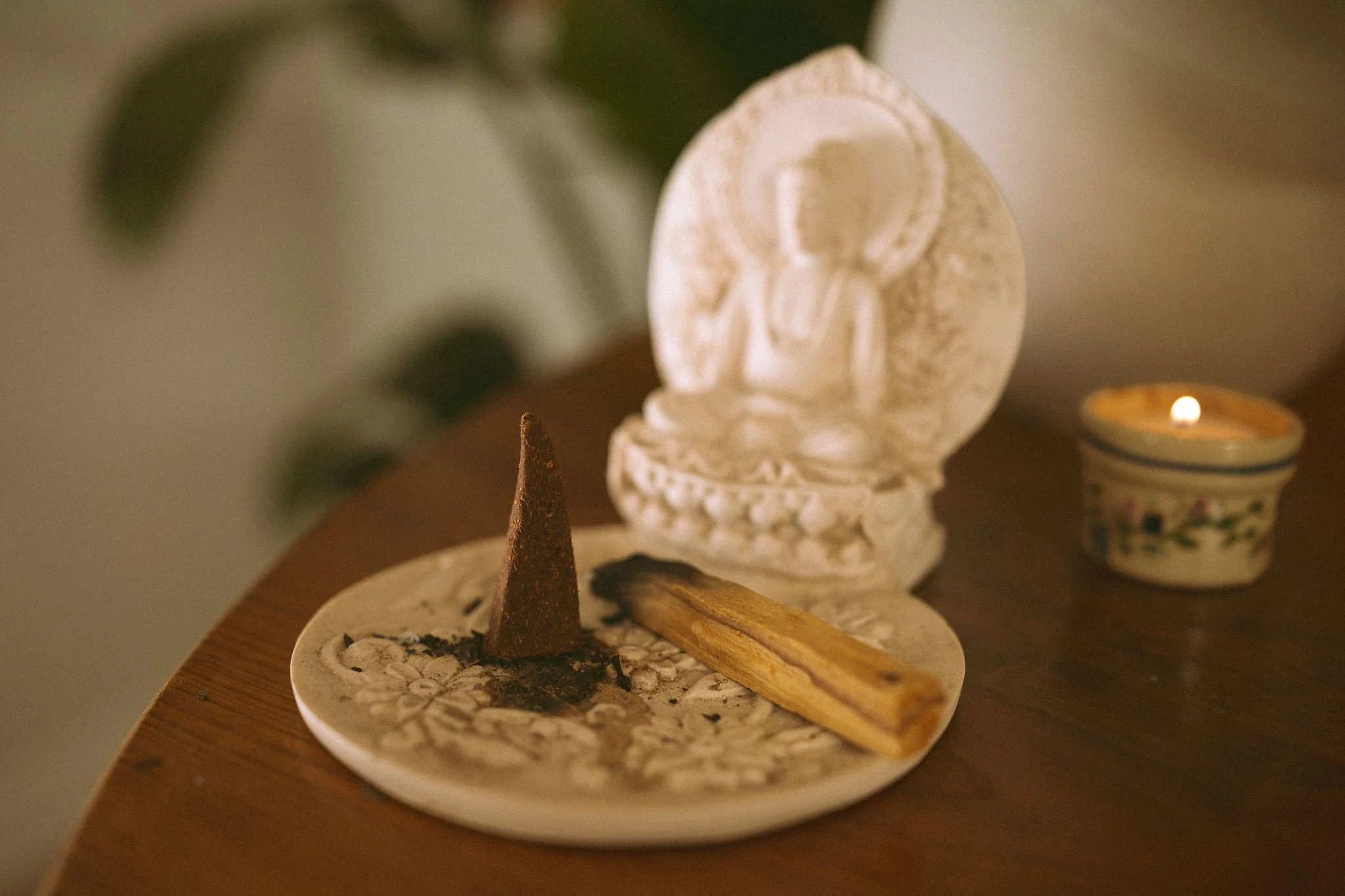A decorative arrangement on a wooden surface includes a white carved angel figurine, a small lit candle in a floral-patterned holder, two sticks of Palo Santo, and a black conical object, all surrounded by small white stones.