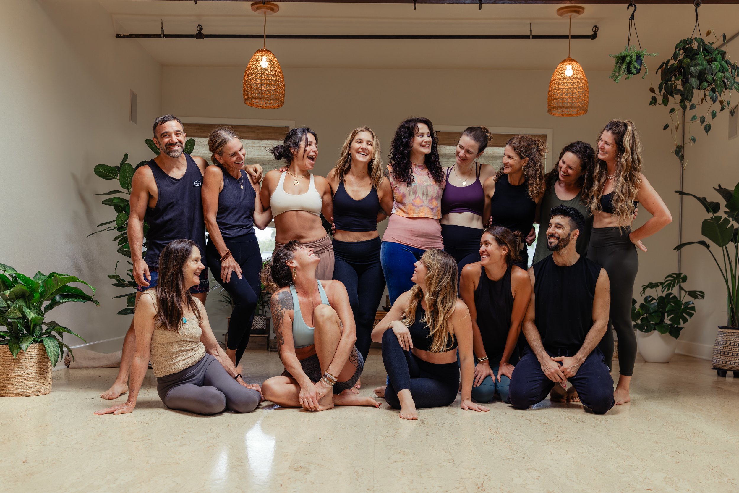 Group of people participating in a yoga or wellness class in a room with plants and hanging lights, some sitting and others standing, smiling and relaxed.