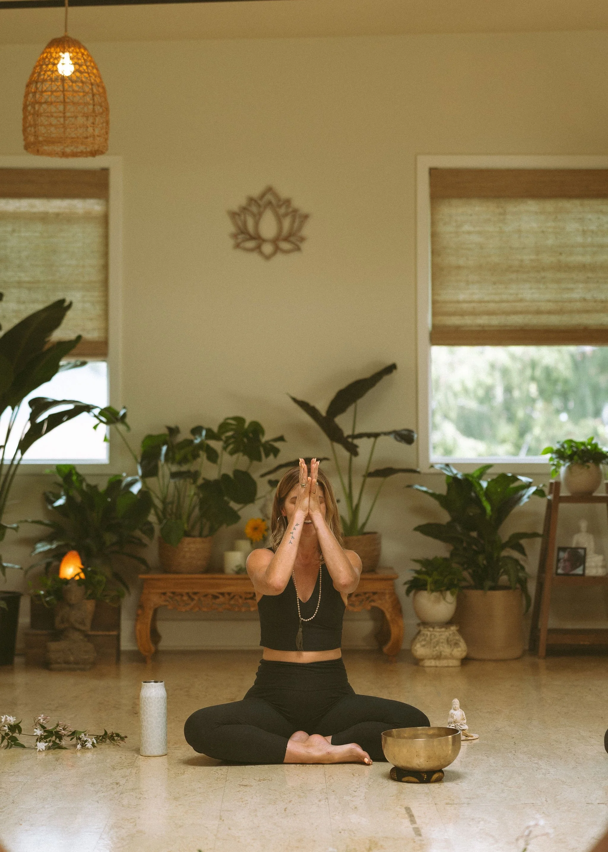 Woman practicing yoga or meditation indoors, sitting cross-legged on the floor, with plants and decorative items in the background.