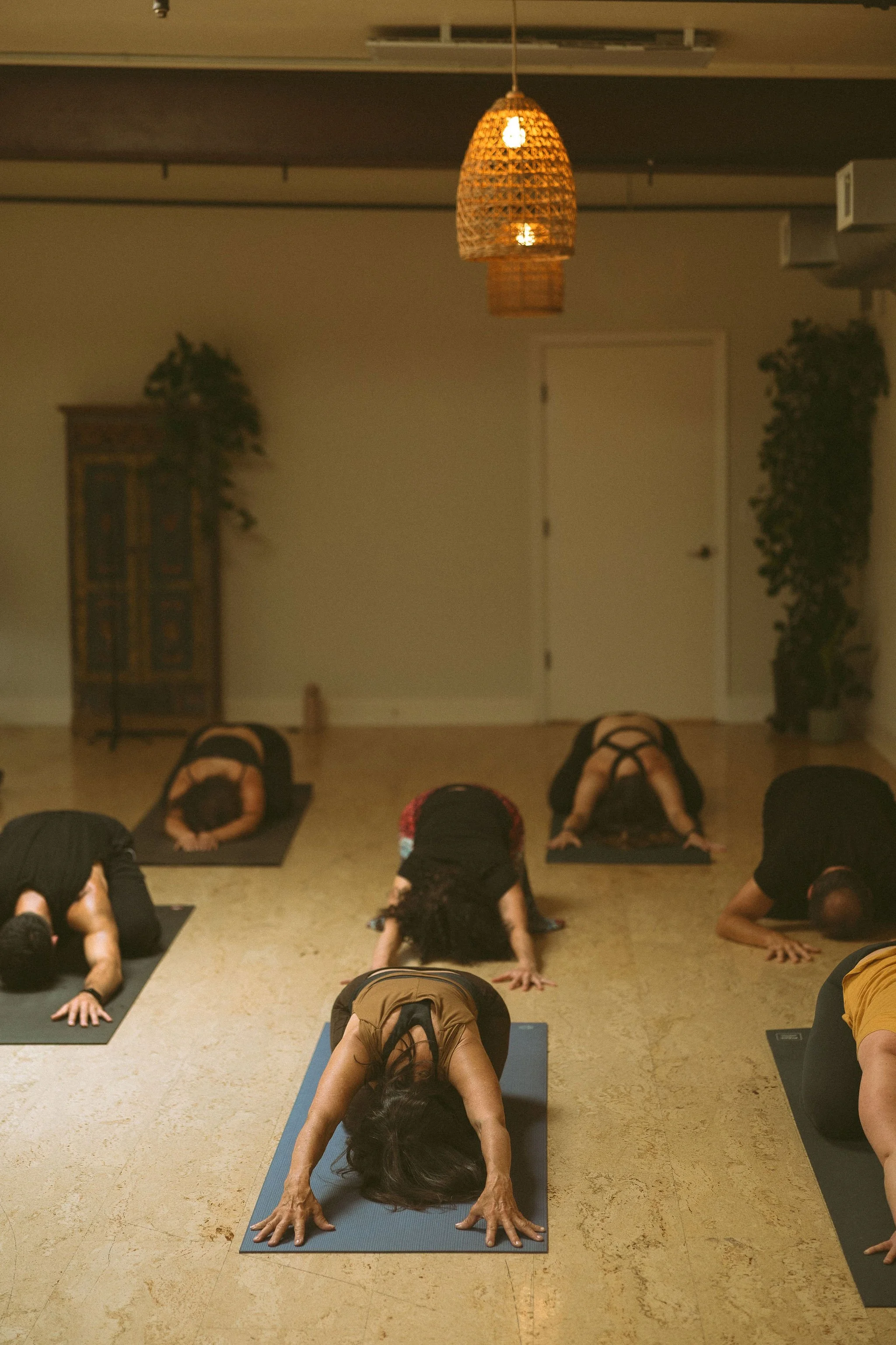 A group of people participating in a yoga class in a room with beige walls. They are in child’s pose on yoga mats, facing downward, with hands extended forward and knees on the floor. The room has warm lighting from a hanging wicker lamp, a wooden ca