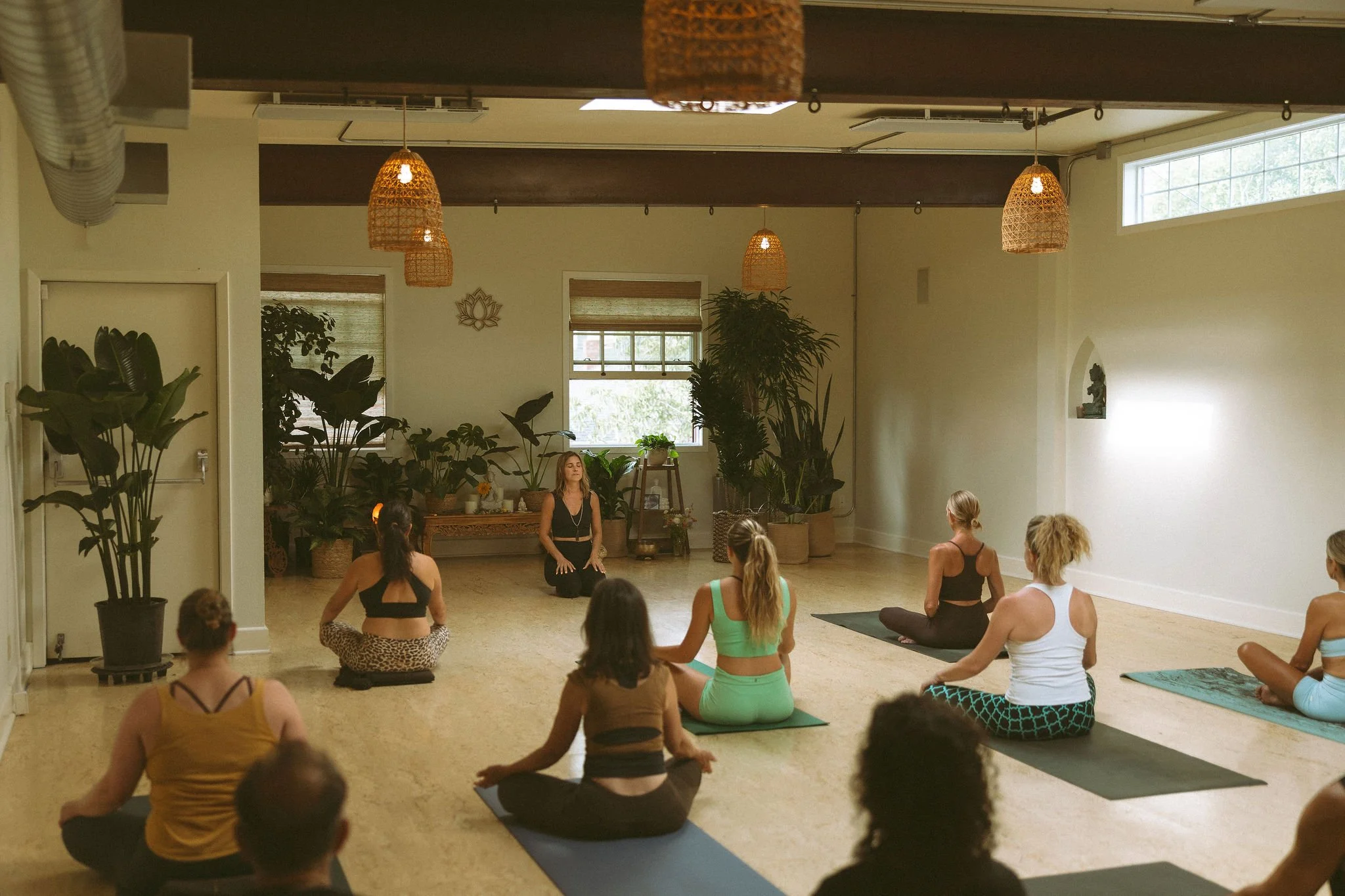 A group of women participating in a yoga or meditation class in a bright, spacious room with plants, hanging wicker lights, and large windows, following an instructor seated in the center.