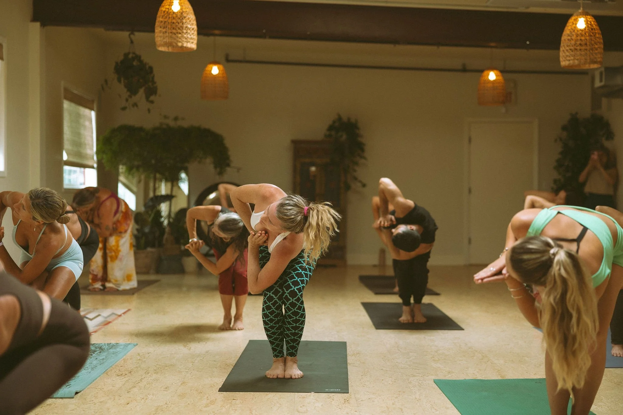 Group of people practicing yoga in a studio, with someParticipants in a forward bend pose and a woman in the center in a prayer pose.