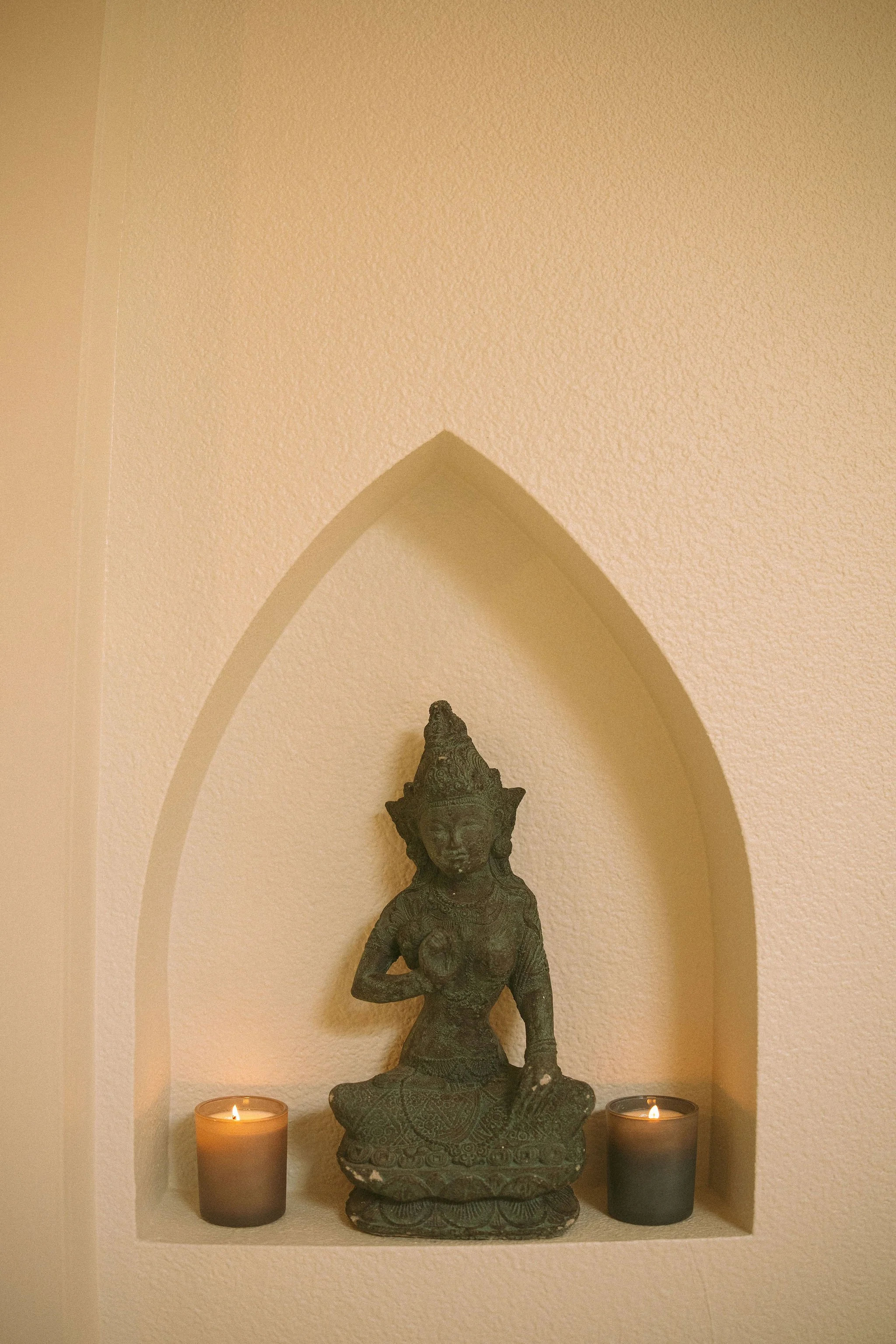 A small shrine recess with a metal statue of a seated deity, flanked by two lit candles, set against a beige wall with a textured finish.