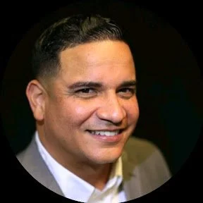 Close-up of a smiling man in a suit with dark hair and a light shirt against a dark background.
