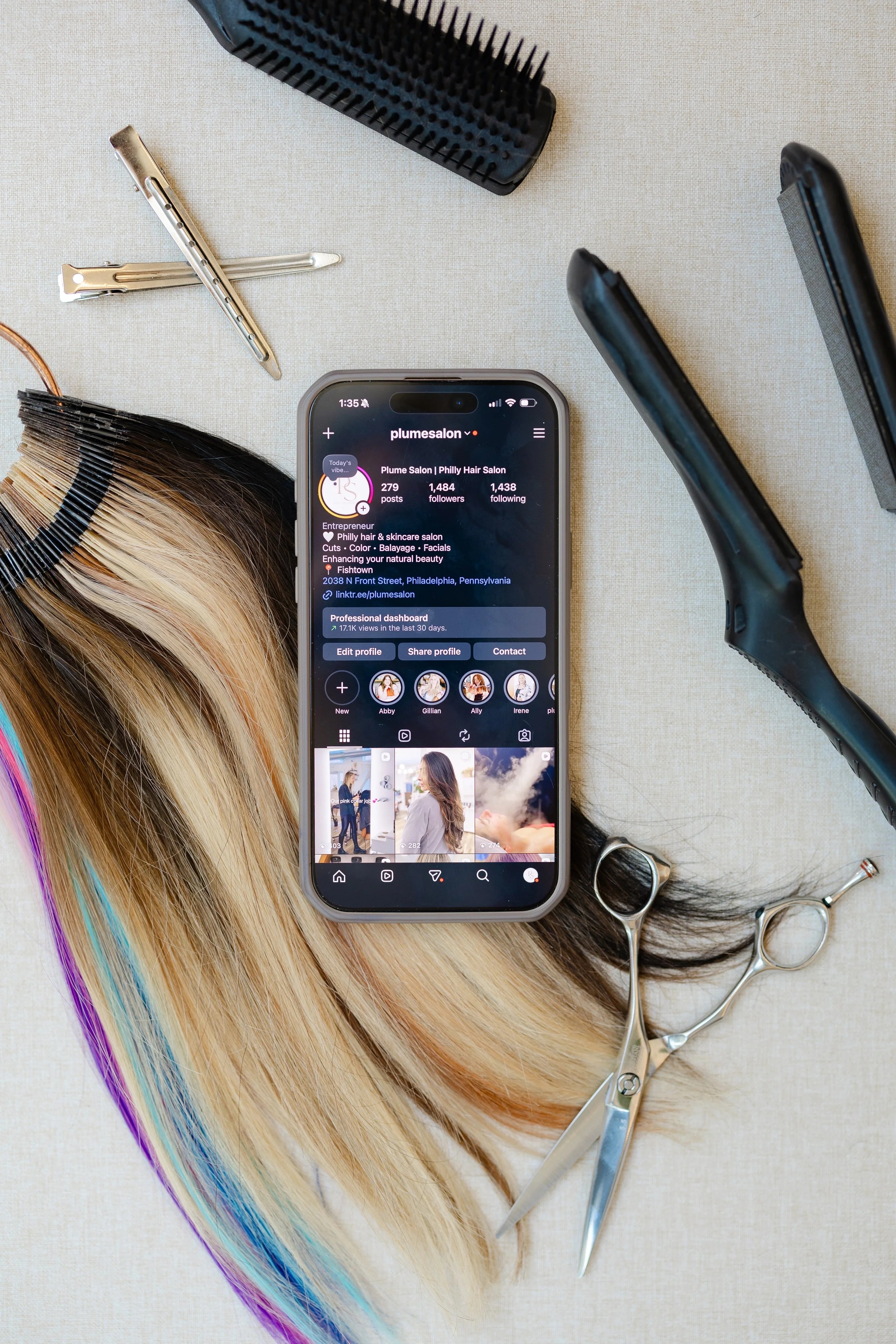 A flat lay of hairdressing tools and accessories on a beige surface. Includes a section of a blonde and rainbow-colored wig, a black hairbrush, two pairs of silver scissors, and a smartphone displaying an Instagram profile for Plume Salon, a Philly hair salon.