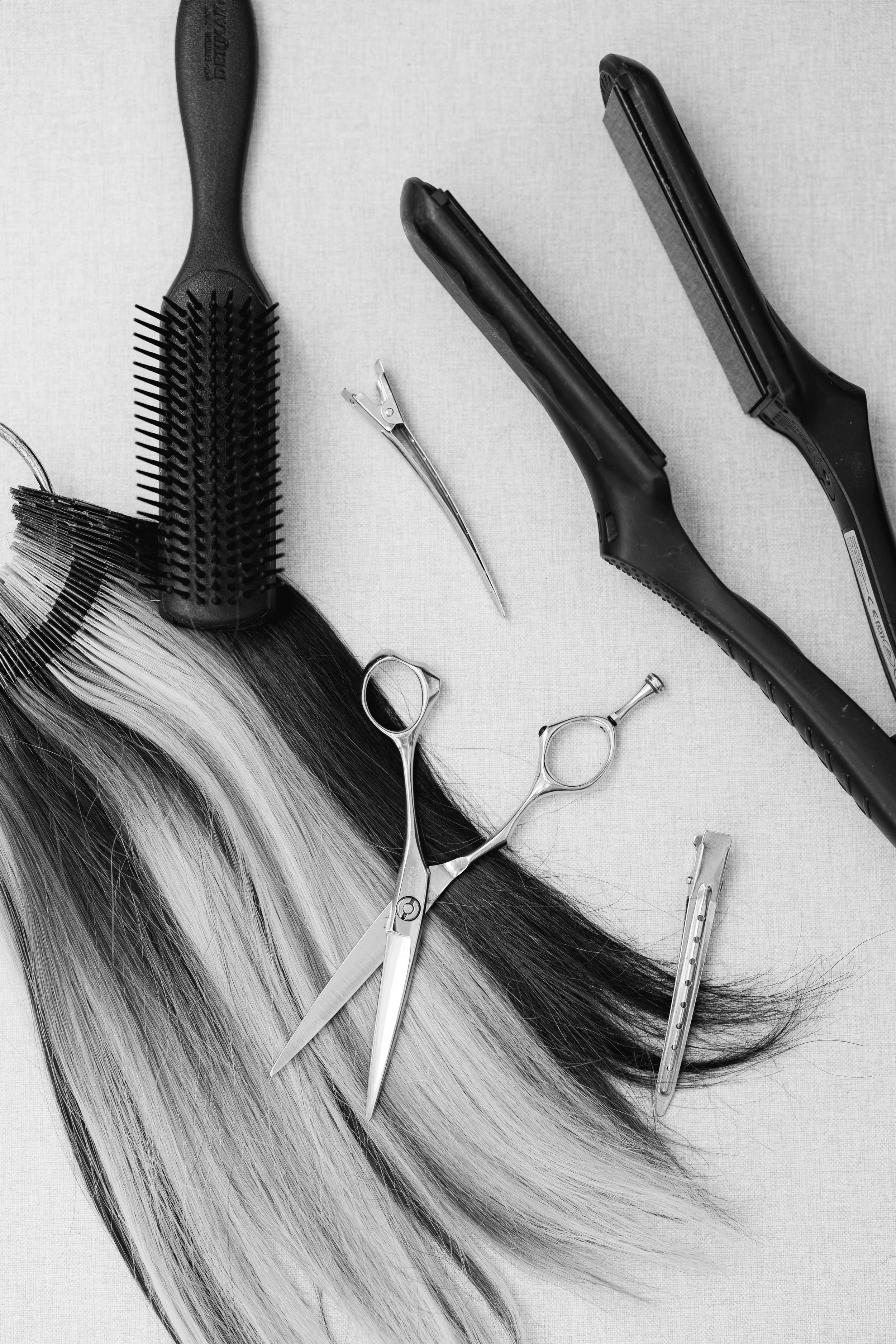 Salon hair styling tools including a round brush, flat irons, scissors, and hair clips arranged on a surface with black and white hair strands.