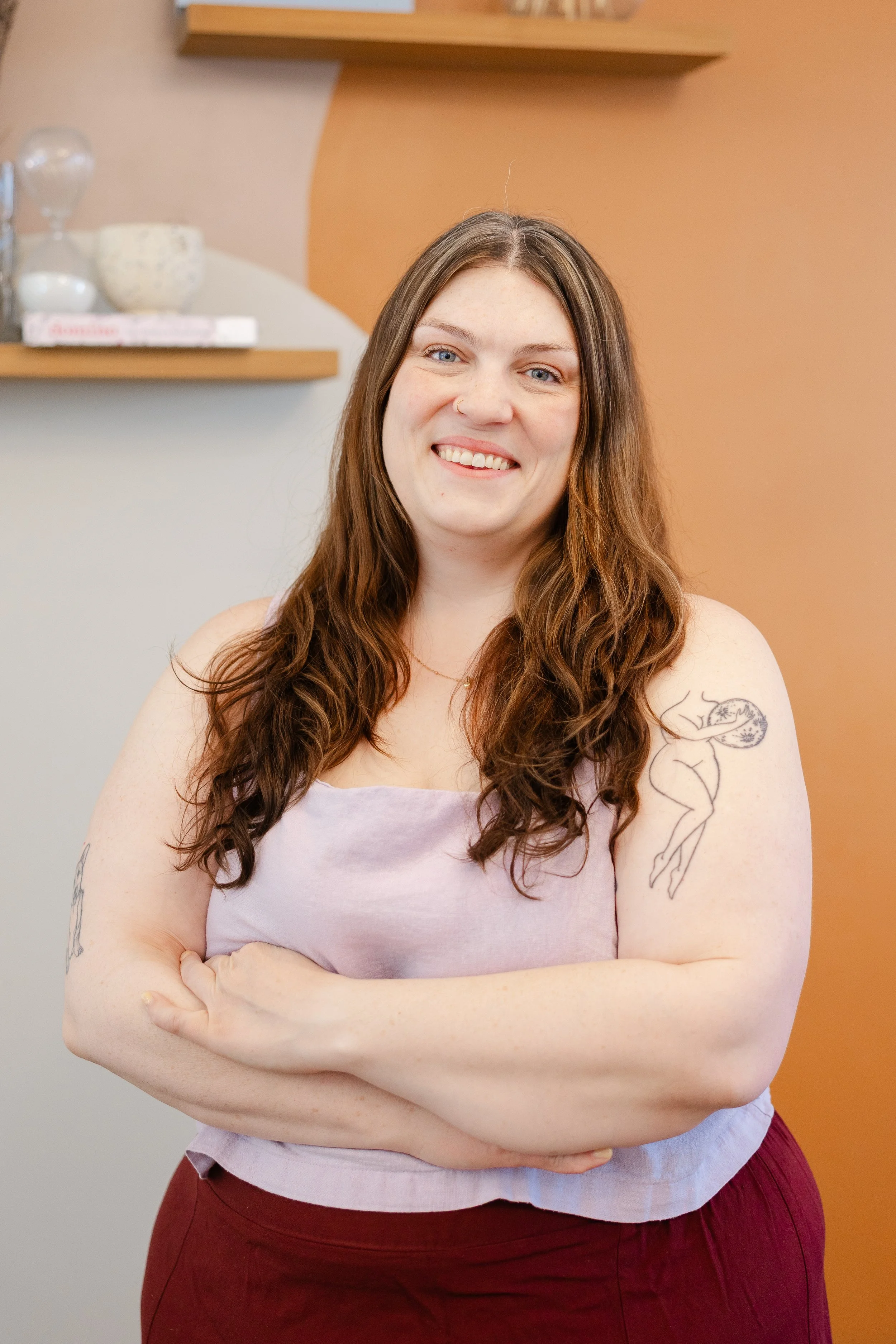 A smiling woman with long, wavy brown hair, tattoos on her arms, wearing a light purple sleeveless top and maroon pants, standing with crossed arms against a colorful background.