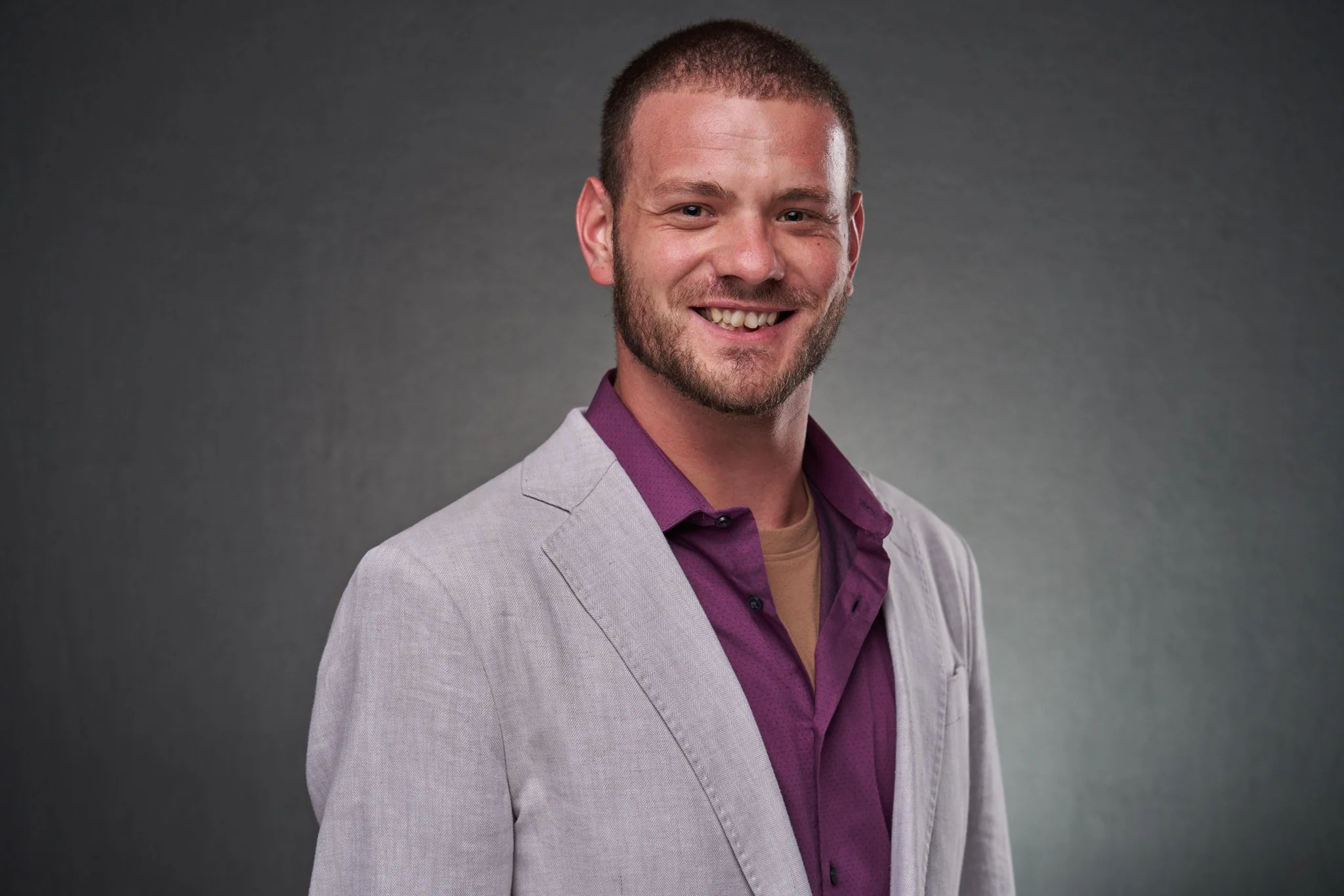 A smiling man with short brown hair and a beard, wearing a light gray blazer over a purple shirt, standing against a dark gray background.
