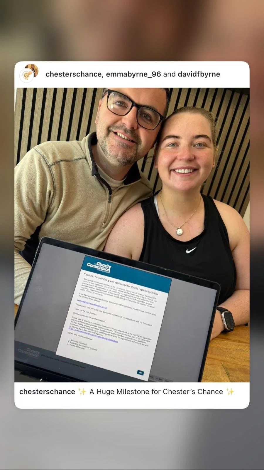 A smiling young woman and an older man are taking a selfie together. The woman is wearing a black sports top and a necklace, and the man is wearing glasses and a beige jacket. They are sitting at a table with a laptop in front of them displaying a ch