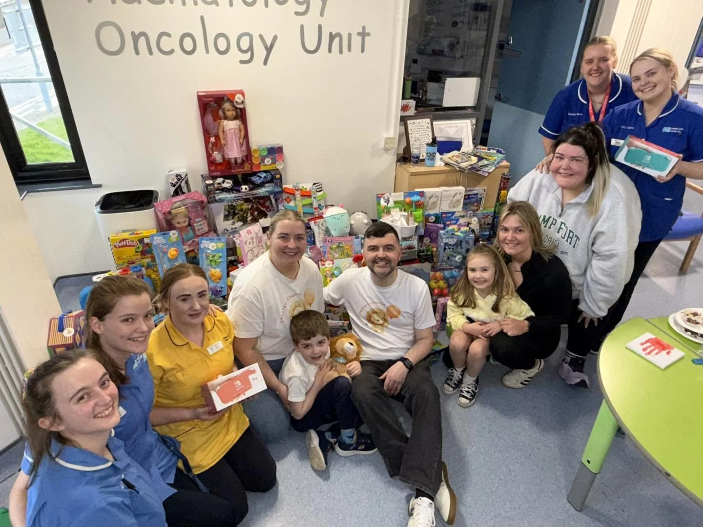 Group of people, including children and adults, gathered around a table with toys, in a hospital room labeled 'Oncology Unit'. Some are holding toys and smiling at the camera.