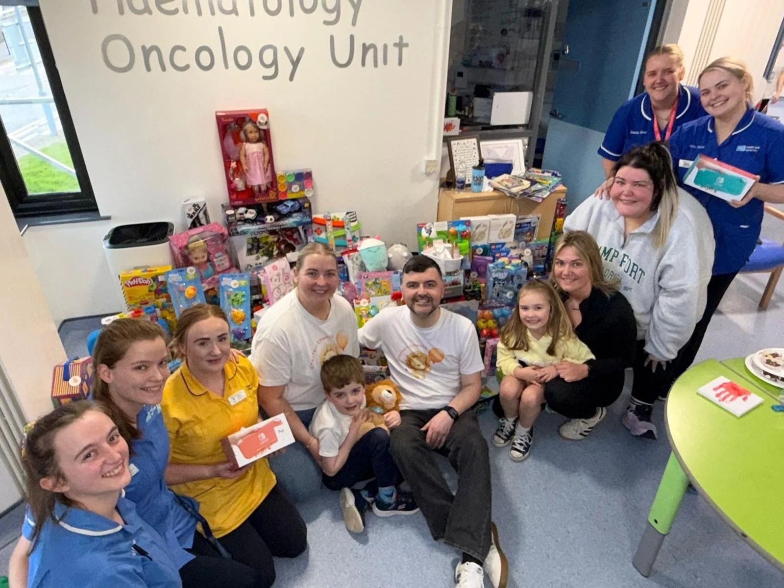 Group of people, including children and adults, gathered in a room with toys, clothes, and books for a donation or charity event at a hospital's Hematology Oncology Unit.