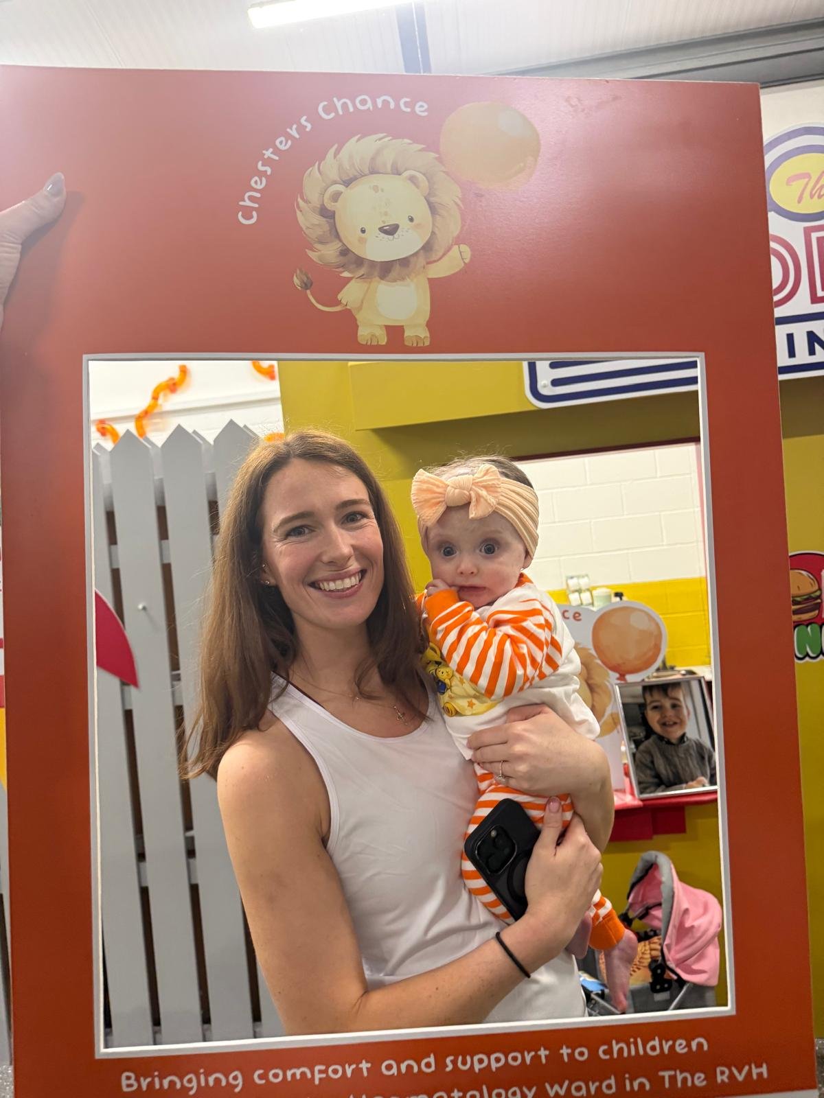 A woman holding a baby in front of a mirror with a decorative frame. The woman is smiling and the baby has a curious expression. The frame features a cartoon lion and the words "Chester's chance" at the top, with colorful background elements.