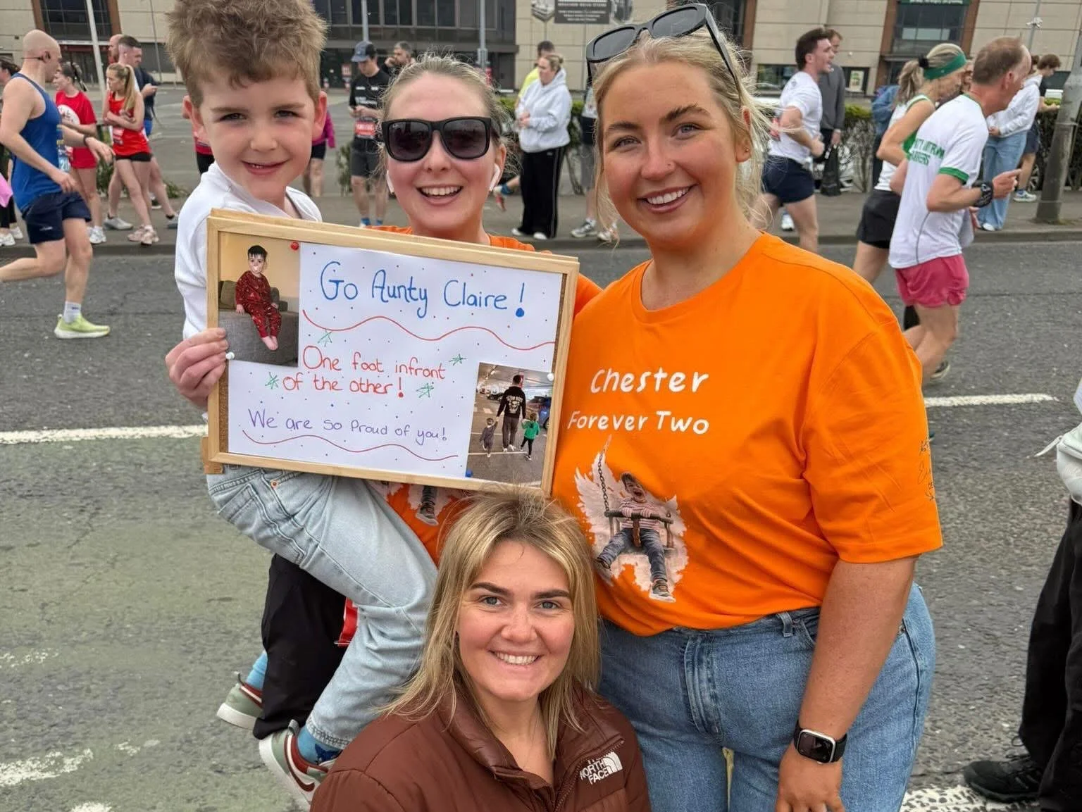 A group of women and a young boy at a running event. One woman is holding a sign supporting a girl named Aunty Claire, with photos and messages, while another woman is kneeling in front. The young boy is sitting on the woman's shoulders, smiling, and