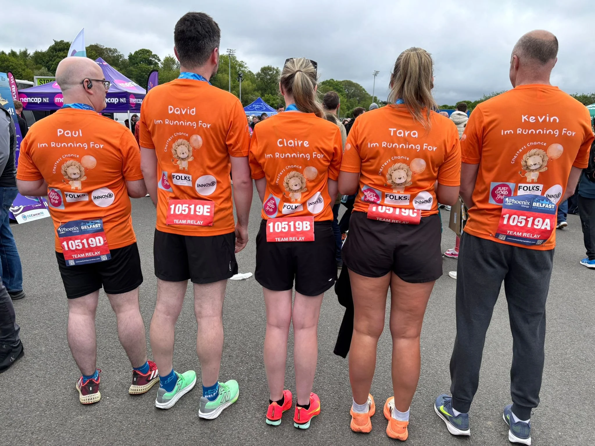 A group of five people, wearing orange shirts with names and running slogans, standing with their backs to the camera at a marathon event, with other runners and event tents in the background.