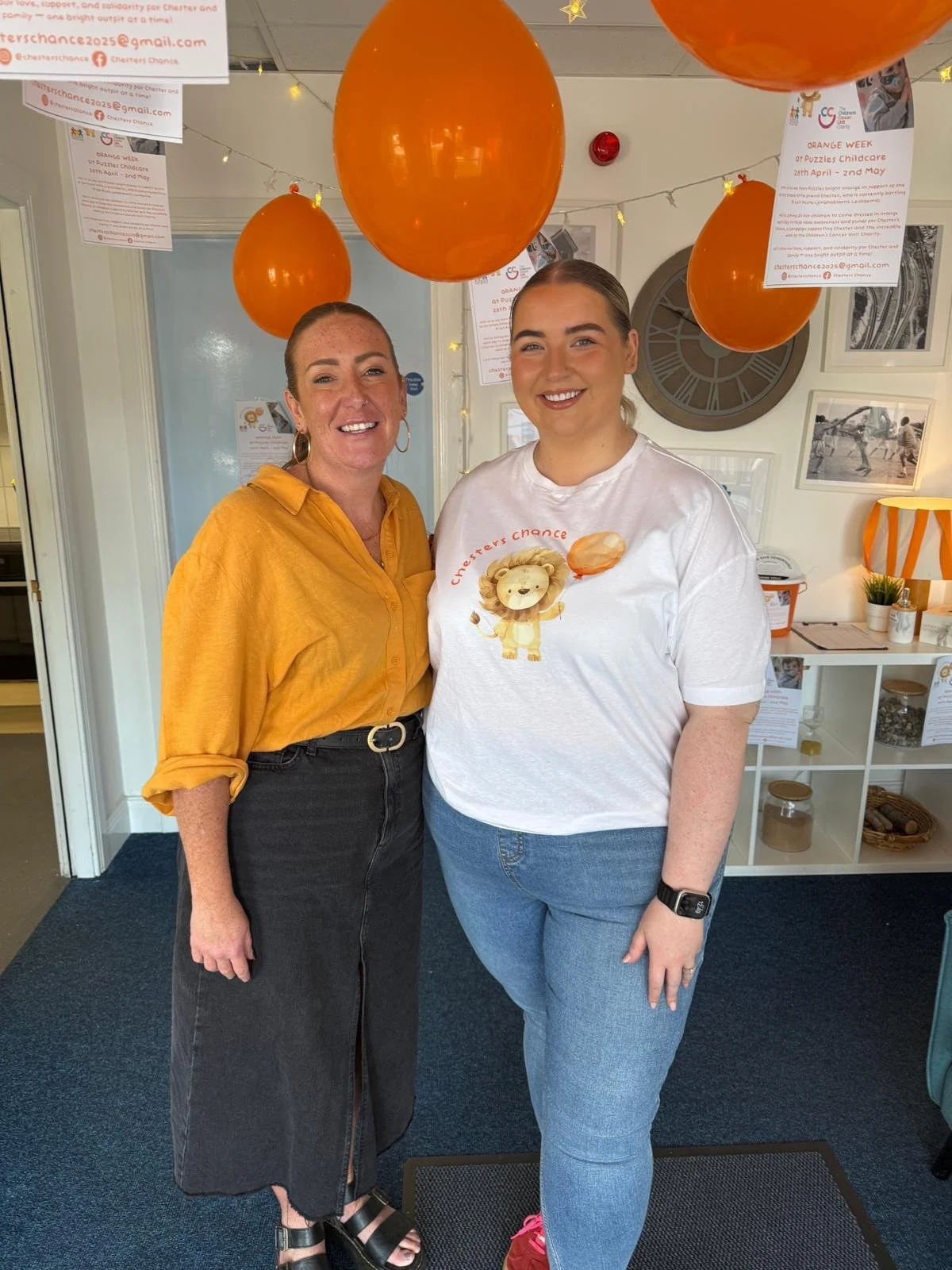 Two women standing inside a room decorated with orange balloons and fairy lights, smiling at the camera. One woman wears a yellow shirt and black skirt, the other wears a white t-shirt with a lion graphic and blue jeans.