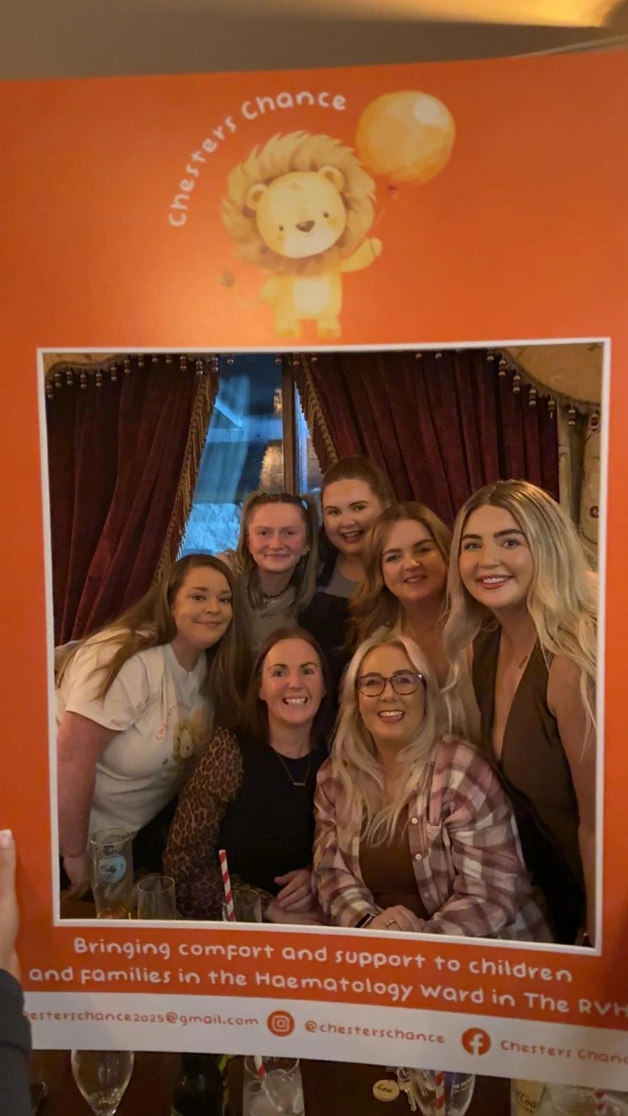 A group of six women smiling and posing for a photo inside a room with red curtains. They are supporting a charity called Chesters Chance, which provides support to children and families in the Hematology ward at The RVH.