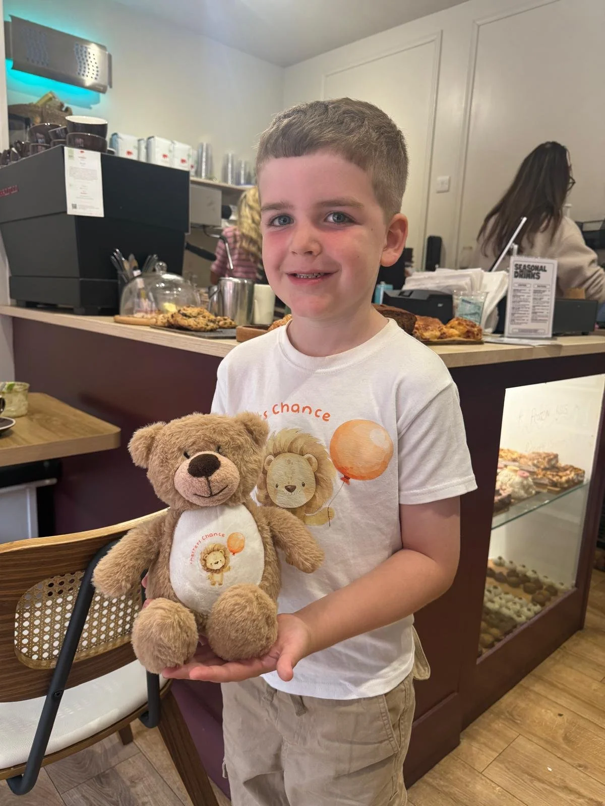 A young boy smiling and holding a teddy bear at a café or bakery. The boy is wearing a white t-shirt with a lion and balloon graphic. The teddy bear has a shirt with a lion and balloon graphic matching the boy's shirt.