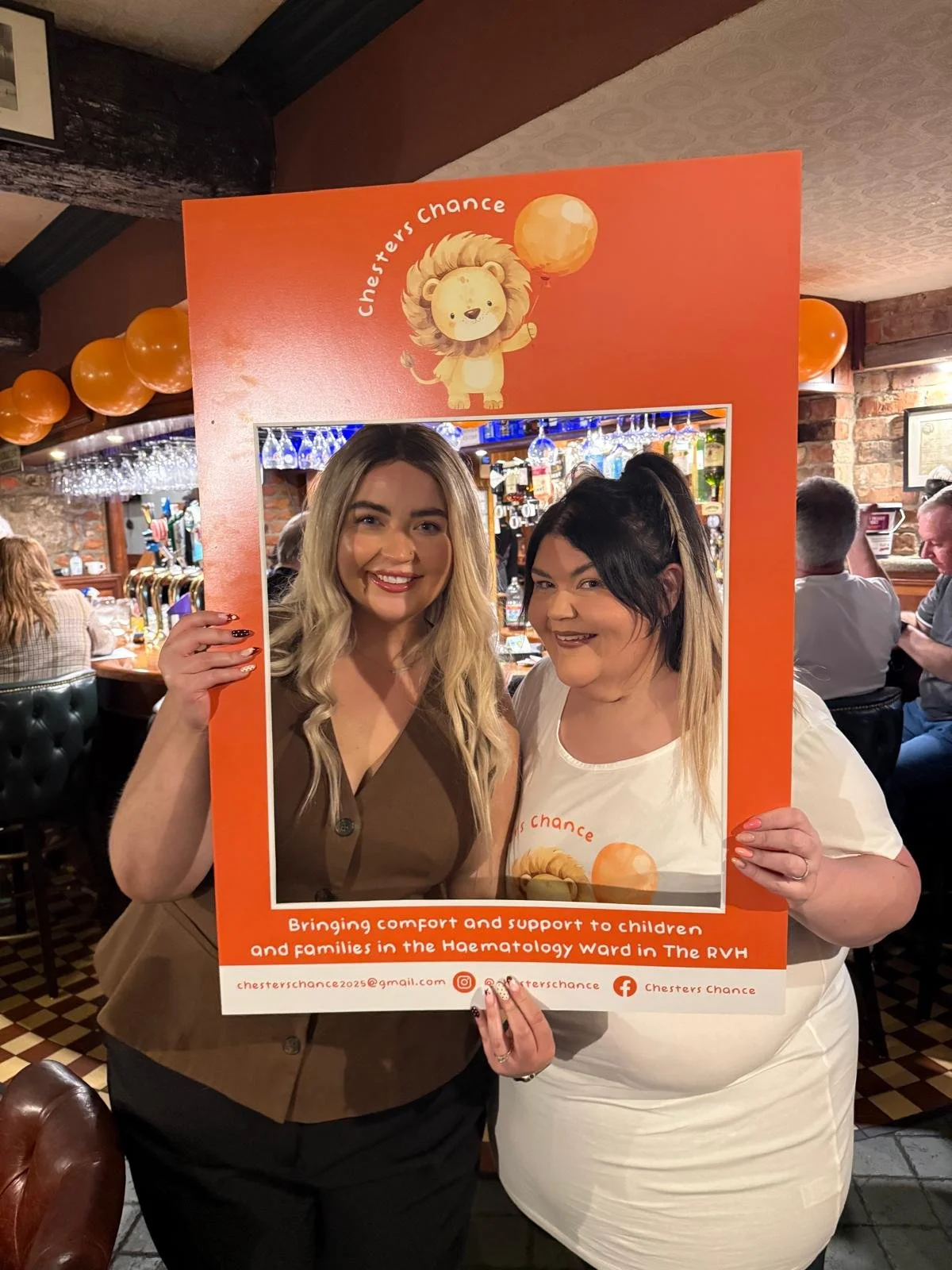 Two smiling women holding a large orange charity photo frame at a bar or pub. The frame features a lion mascot holding a balloon with the text 'Chef's Chance' and a message about supporting children and families in the haemotology ward of The RVH. Th