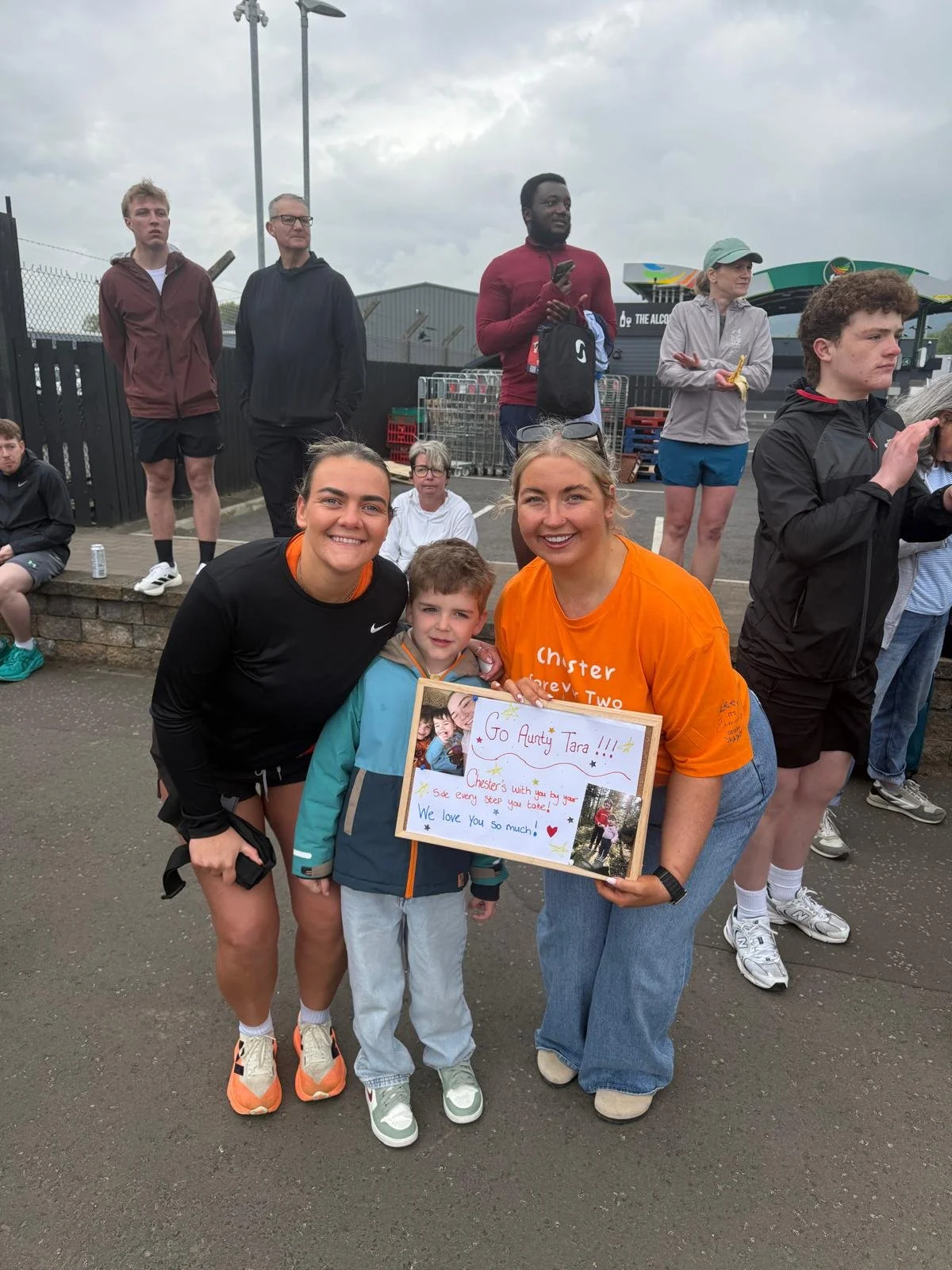 Group of smiling people, including children and adults, at an outdoor event, with one woman holding a framed note that says 'Go Aunty Tara!!!... We love you so much!'