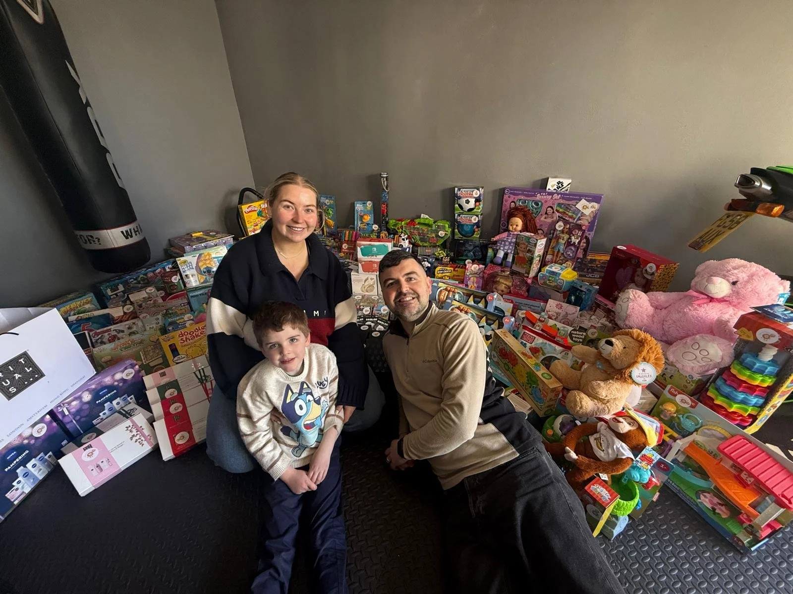 A family of three smiling and sitting among a large collection of toys, including plush animals, dolls, and boxed toys, in a room with plain walls.