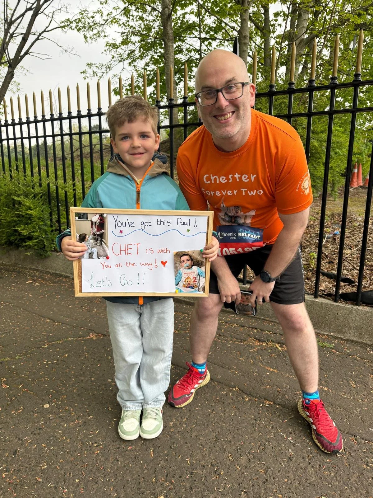 A young boy and an adult man outdoors near a black fence with green trees in the background. The boy holds a decorated sign with photos and writing, and both are smiling.