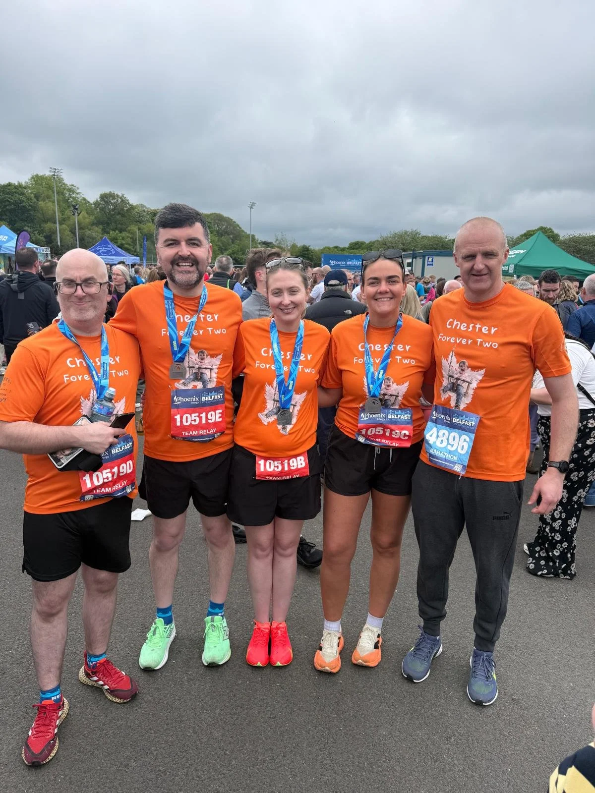 Five runners wearing orange shirts with 'Chester Forever Two' and a graphic print, standing together with medals around their necks at a marathon event, with a crowd and tents in the background on a cloudy day.