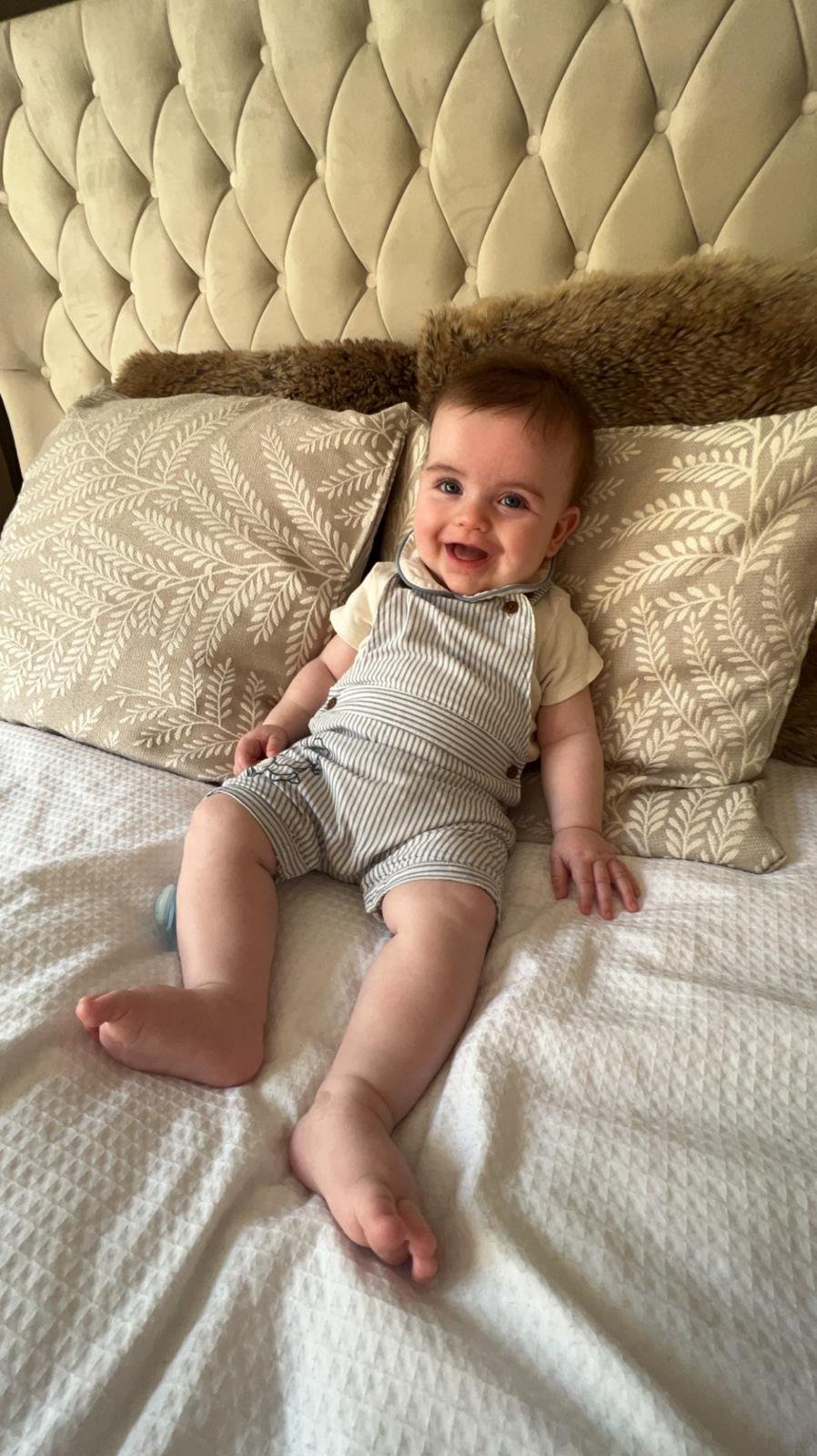 A smiling baby boy lying on a bed with light-colored linens and patterned pillows, holding a relaxed pose.