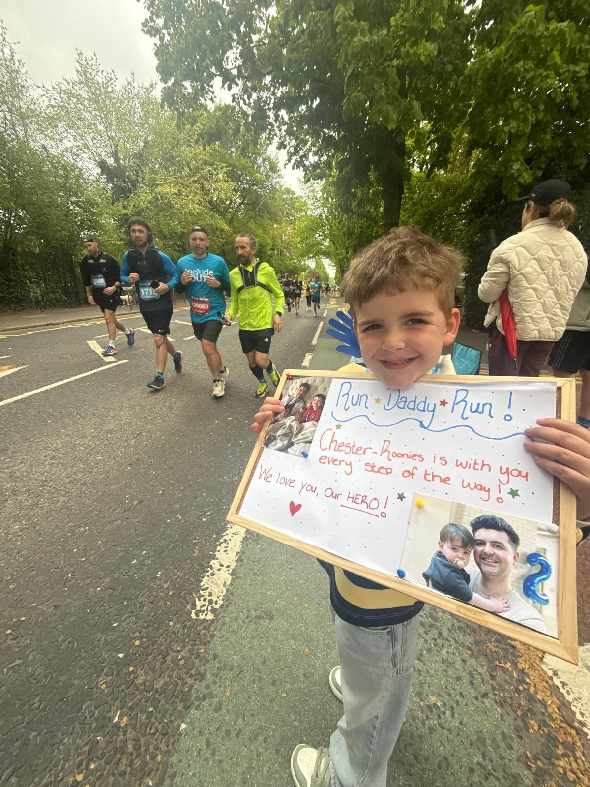 A young boy with a big smile holding a handmade sign during a running event. The sign features a photo of a man and a young child, and a message supporting Chester Roonies, with colorful decorations and a handwritten note.