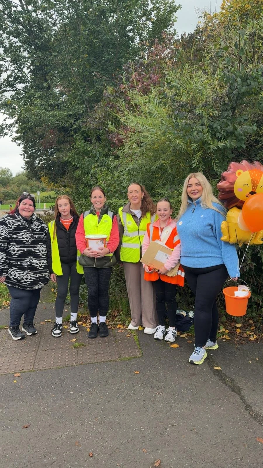Group of six women standing outdoors on a sidewalk, with three wearing high-visibility vests and holding buckets or packages, one holding balloons with a lion and a pumpkin design, in front of green trees and bushes.