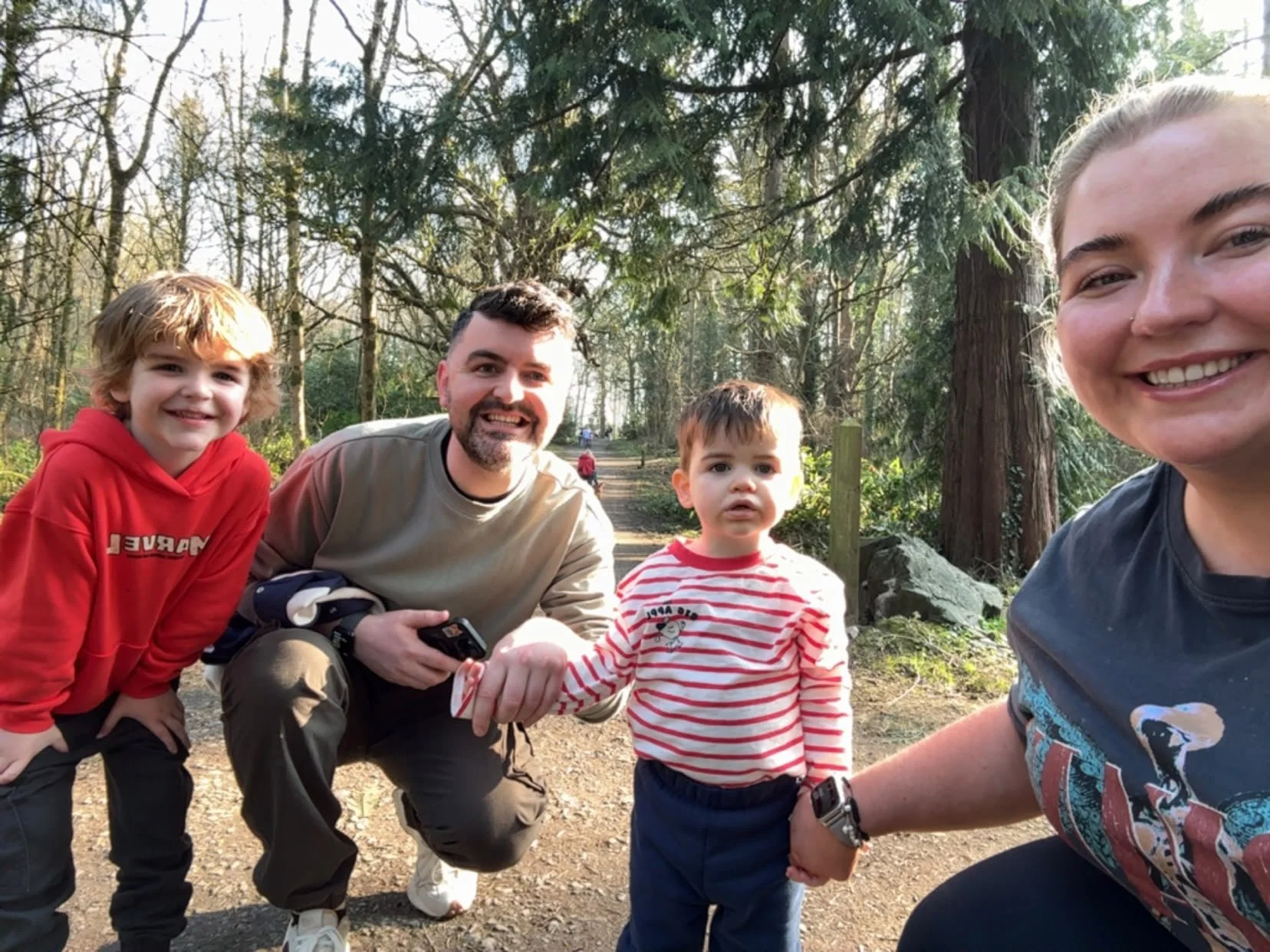 Group of four people in a forested area, people smiling and holding hands, with trees and a dirt path in the background.