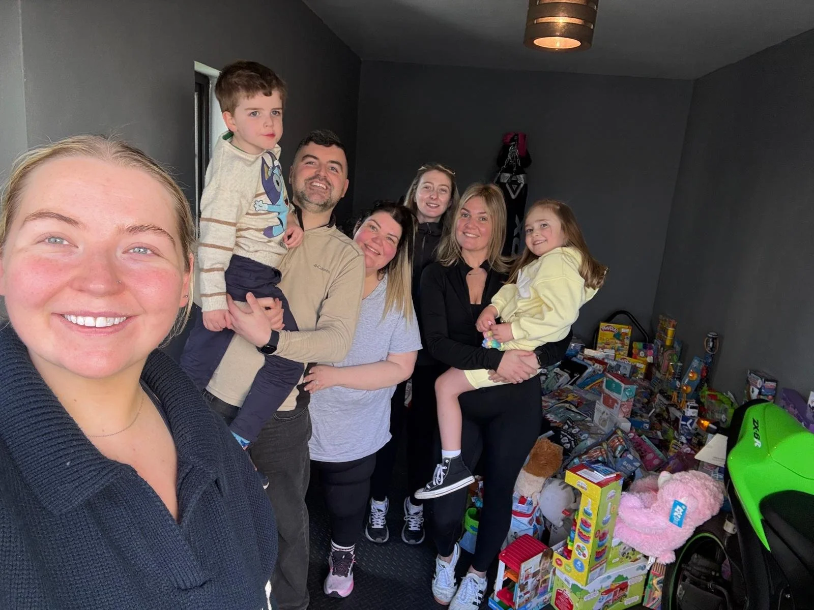A group of seven people standing inside a room with a pile of toys on the floor to the right. The group includes three women, two men, and two young children. They are smiling and posing for the photo.