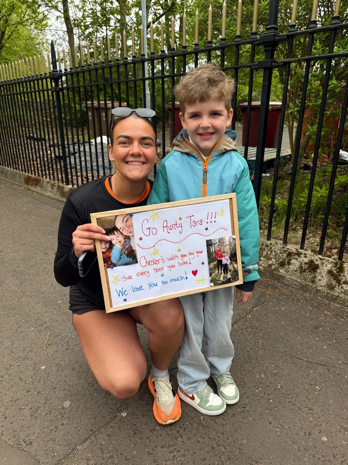 A woman crouching on the sidewalk holding a whiteboard with a message and photos, smiling, and a young boy standing next to her also smiling, in front of a black iron fence with greenery behind. The woman is wearing a black shirt, sunglasses on her h