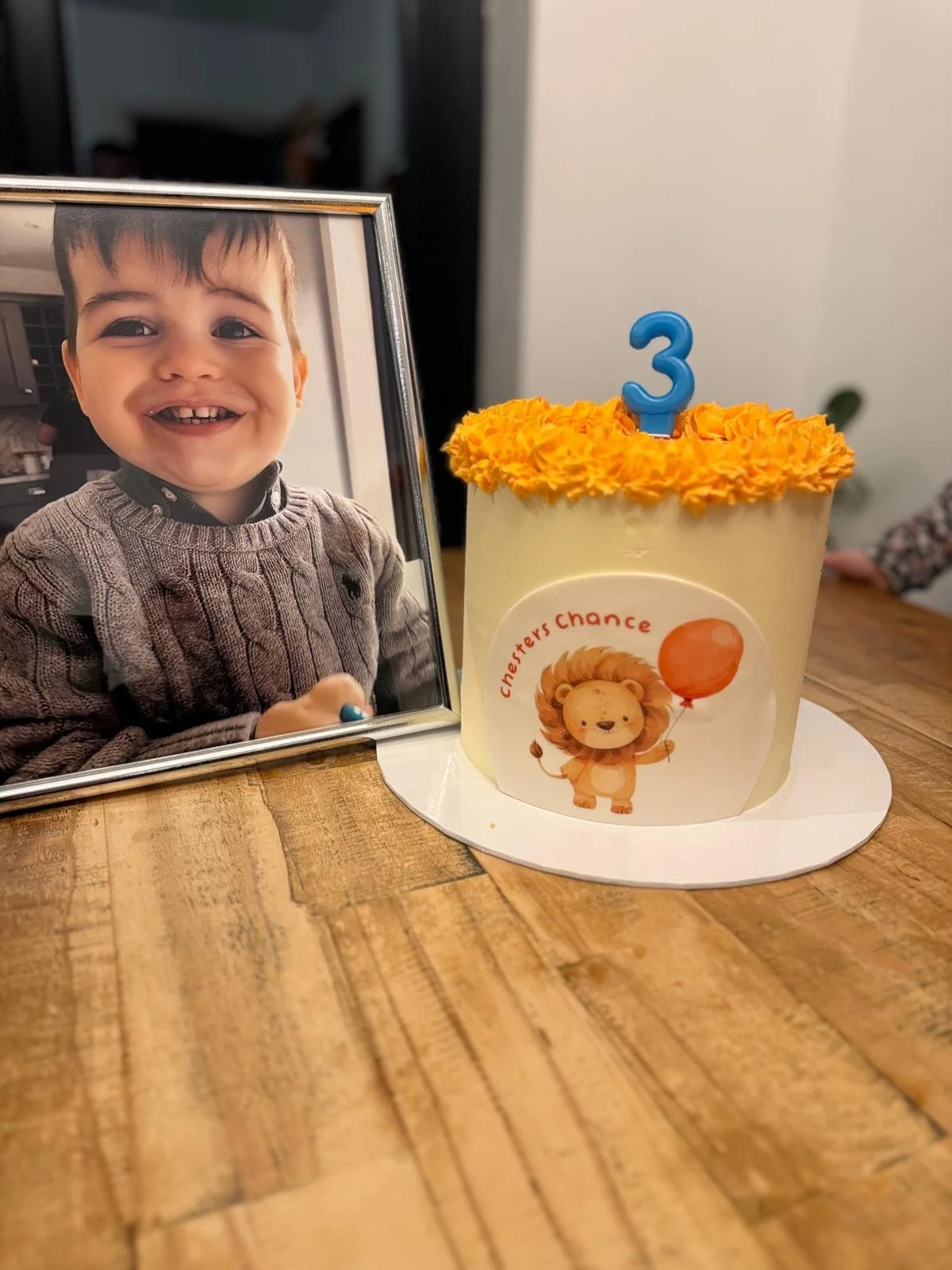 A framed photo of a young boy with a big smile, holding a small object, placed beside a birthday cake with orange decorations and a blue number 3 candle, on a wooden table.