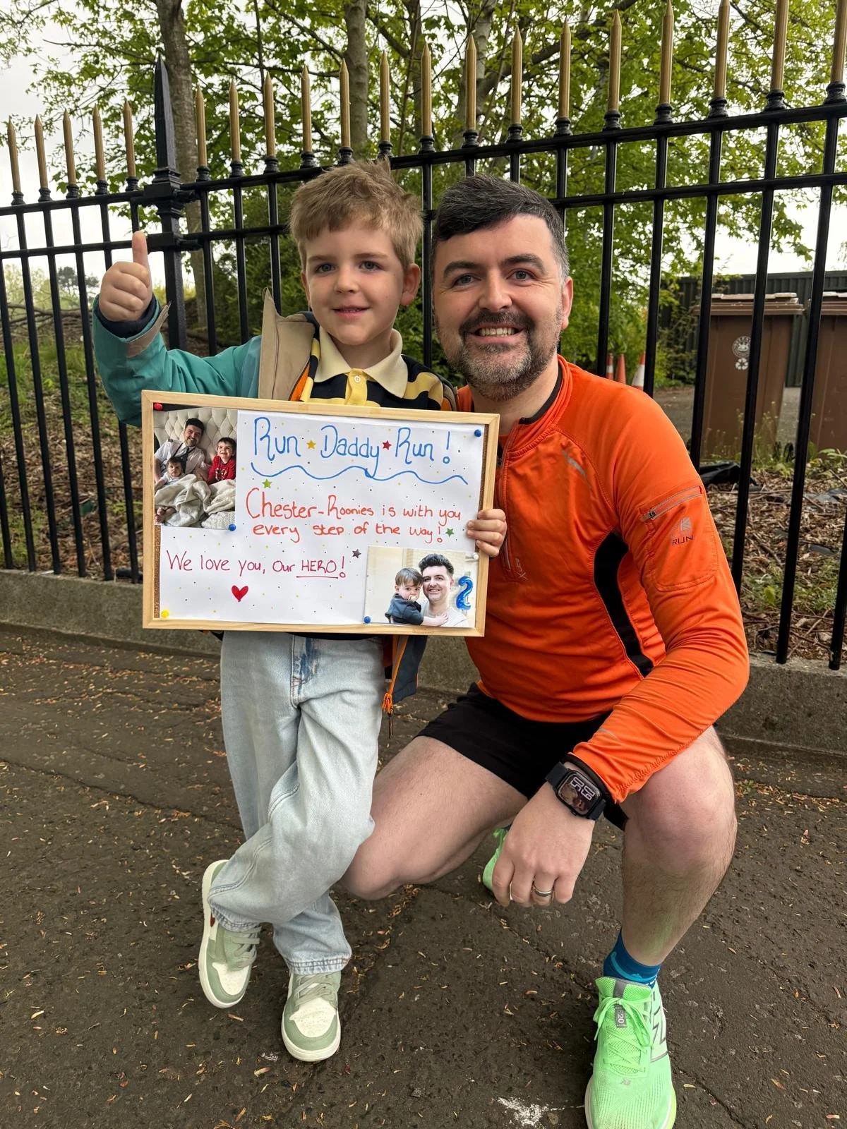 A man and a young boy are outside in front of a black fence. The boy is holding a whiteboard with a message and photos, and giving a thumbs-up. The man is kneeling next to the boy, smiling. The boy is wearing a beige jacket, yellow and black striped 